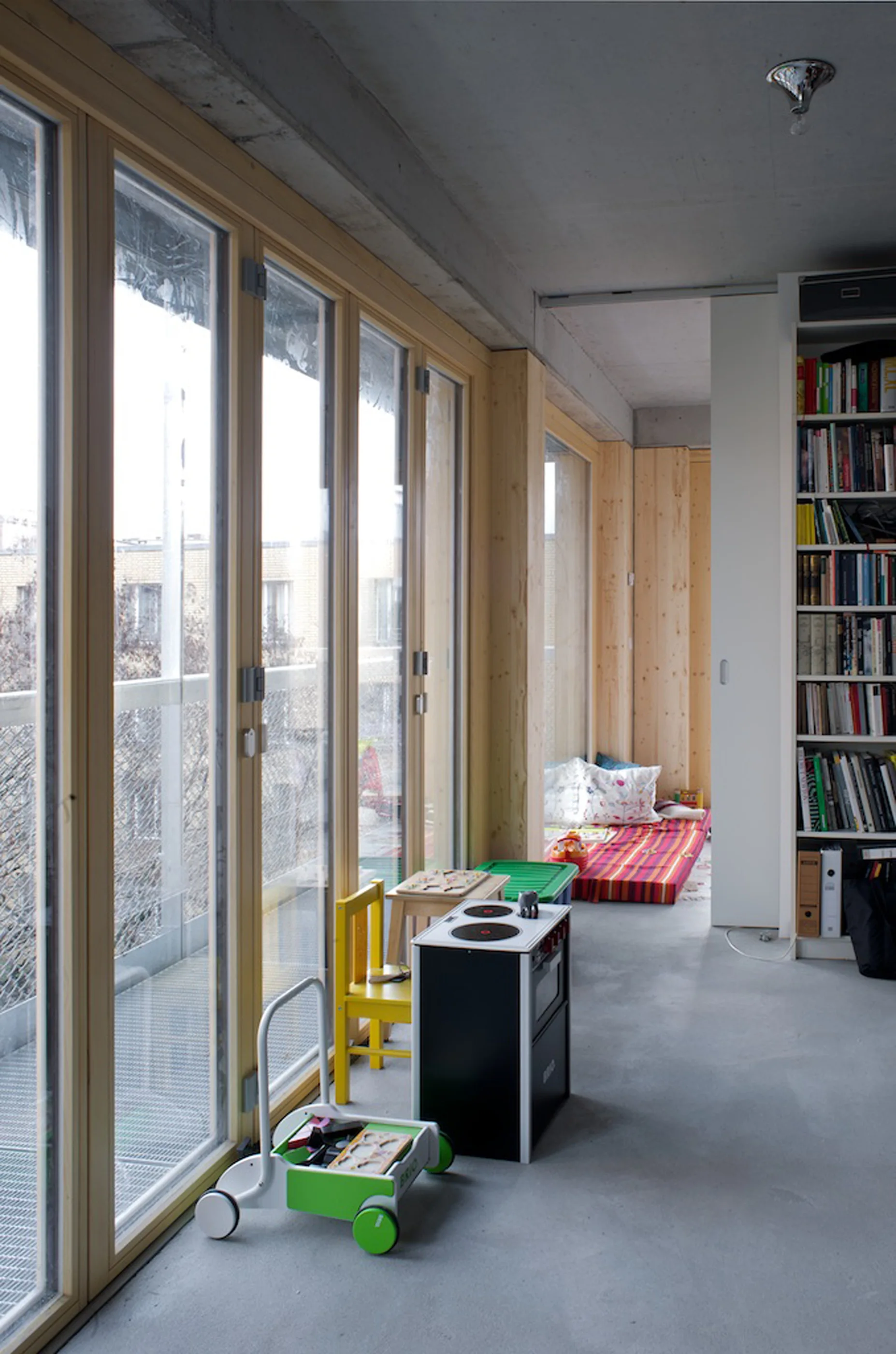 A modern room with an exposed concrete ceiling features floor-to-ceiling wood-framed windows, toys on a grey floor, and a tall white bookcase.