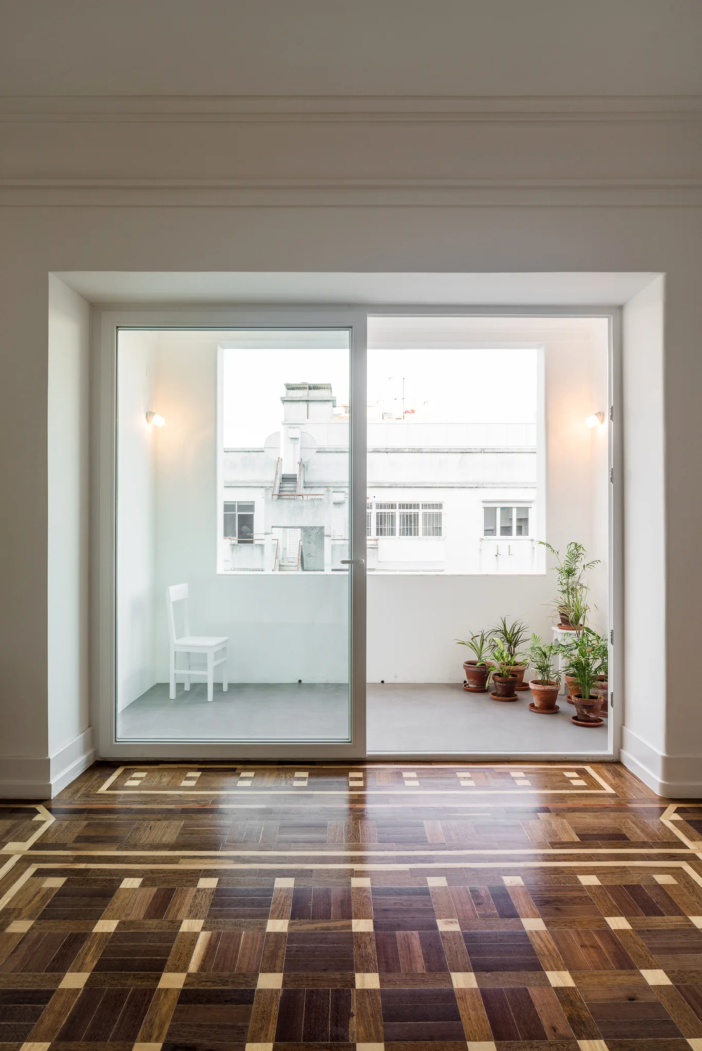 A view through a sliding glass door of a white balcony with potted plants and a white chair, framed by intricate geometric parquet wood flooring.