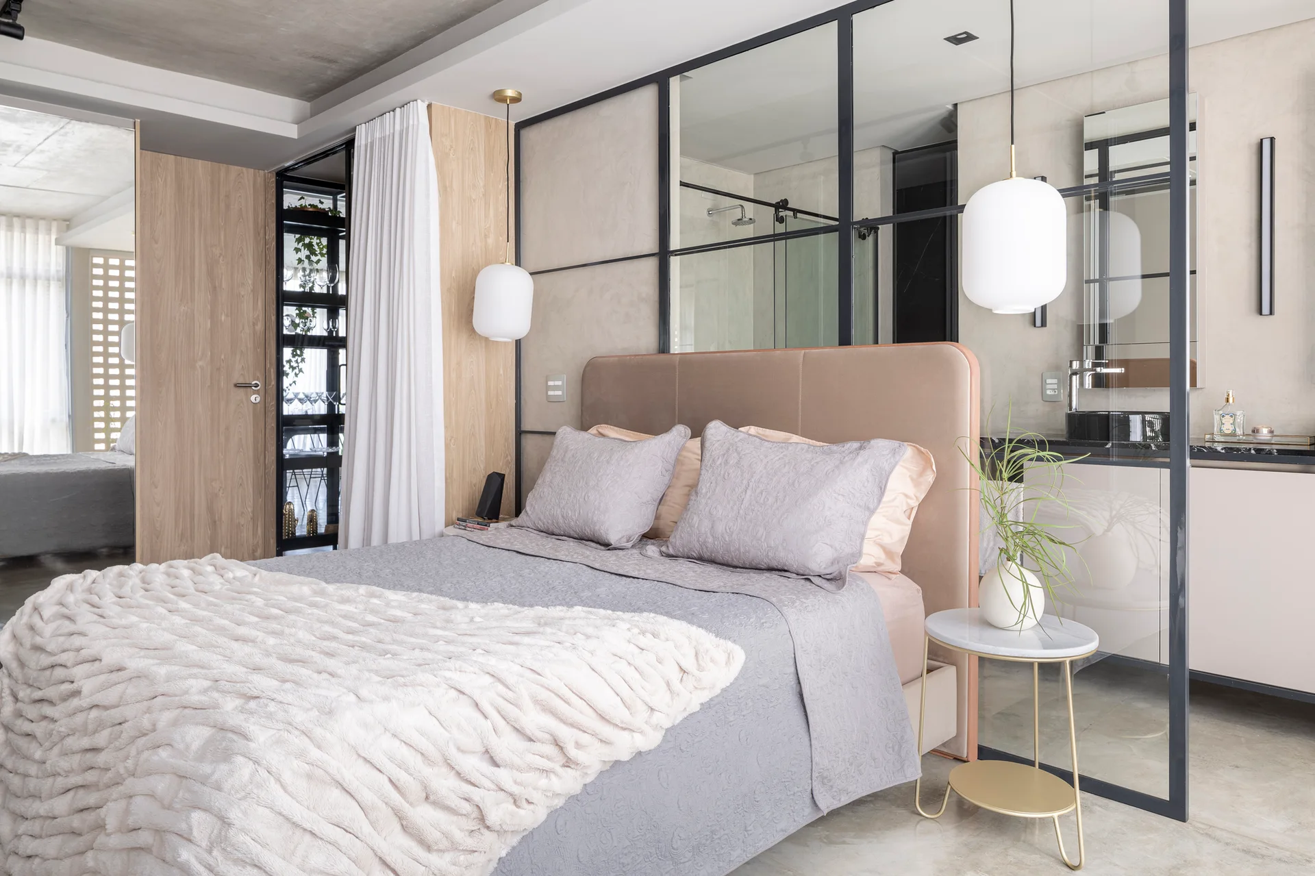 A modern bedroom with an upholstered mauve headboard is separated from an adjacent bathroom by a black-framed glass partition.