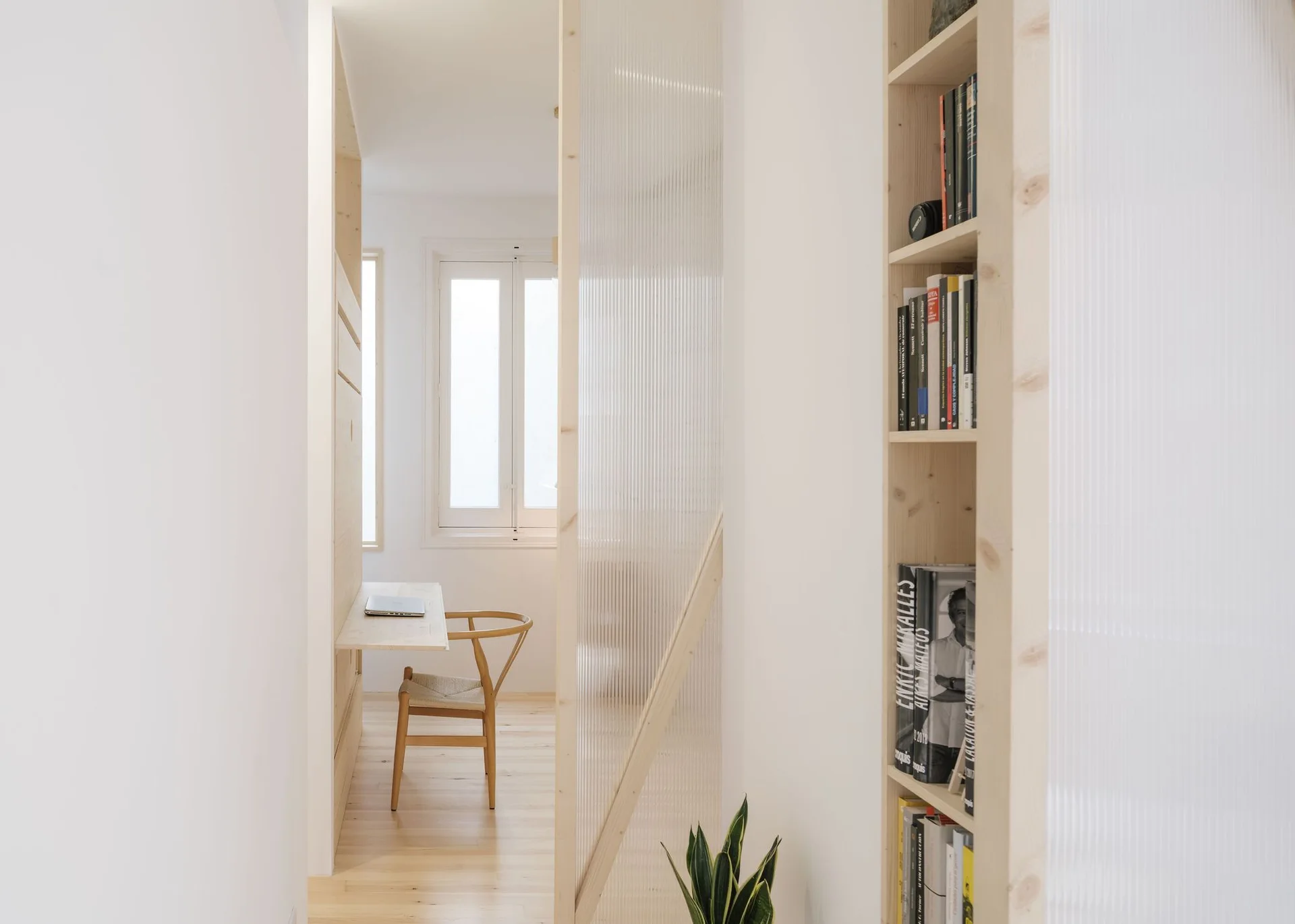 A minimalist home office featuring a light wood wall-mounted desk, built-in shelving, and a tall fluted glass partition panel.