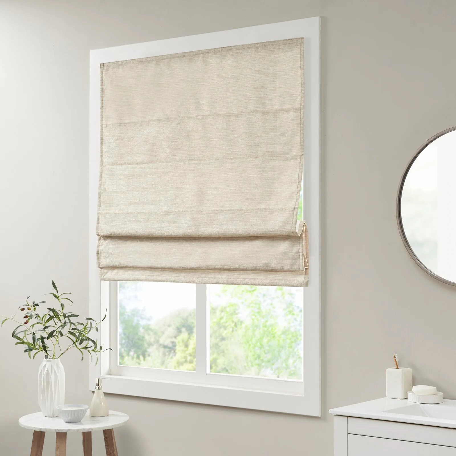 A textured beige fabric Roman shade covers the top portion of a white-framed window next to a bathroom vanity and round mirror.