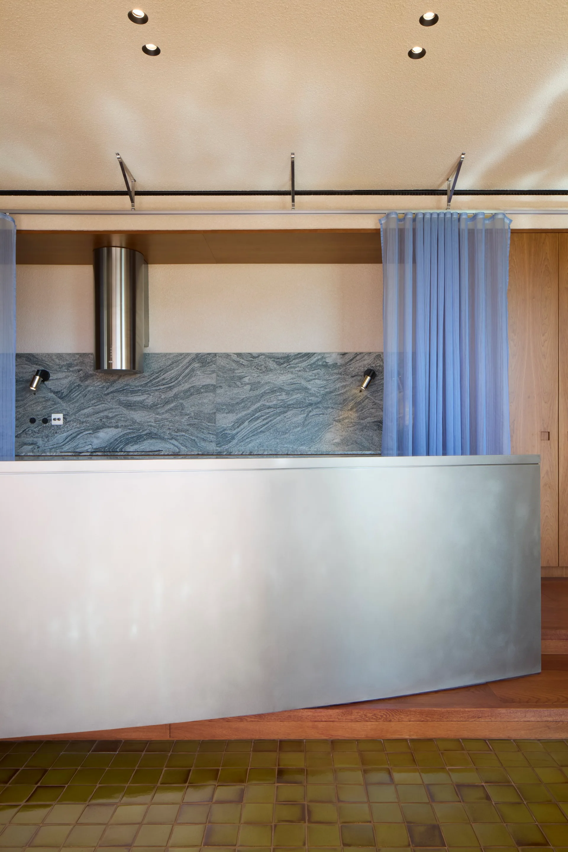 A modern kitchen features a large stainless steel island, a grey veined stone backsplash, wood paneling, and a sheer blue curtain.