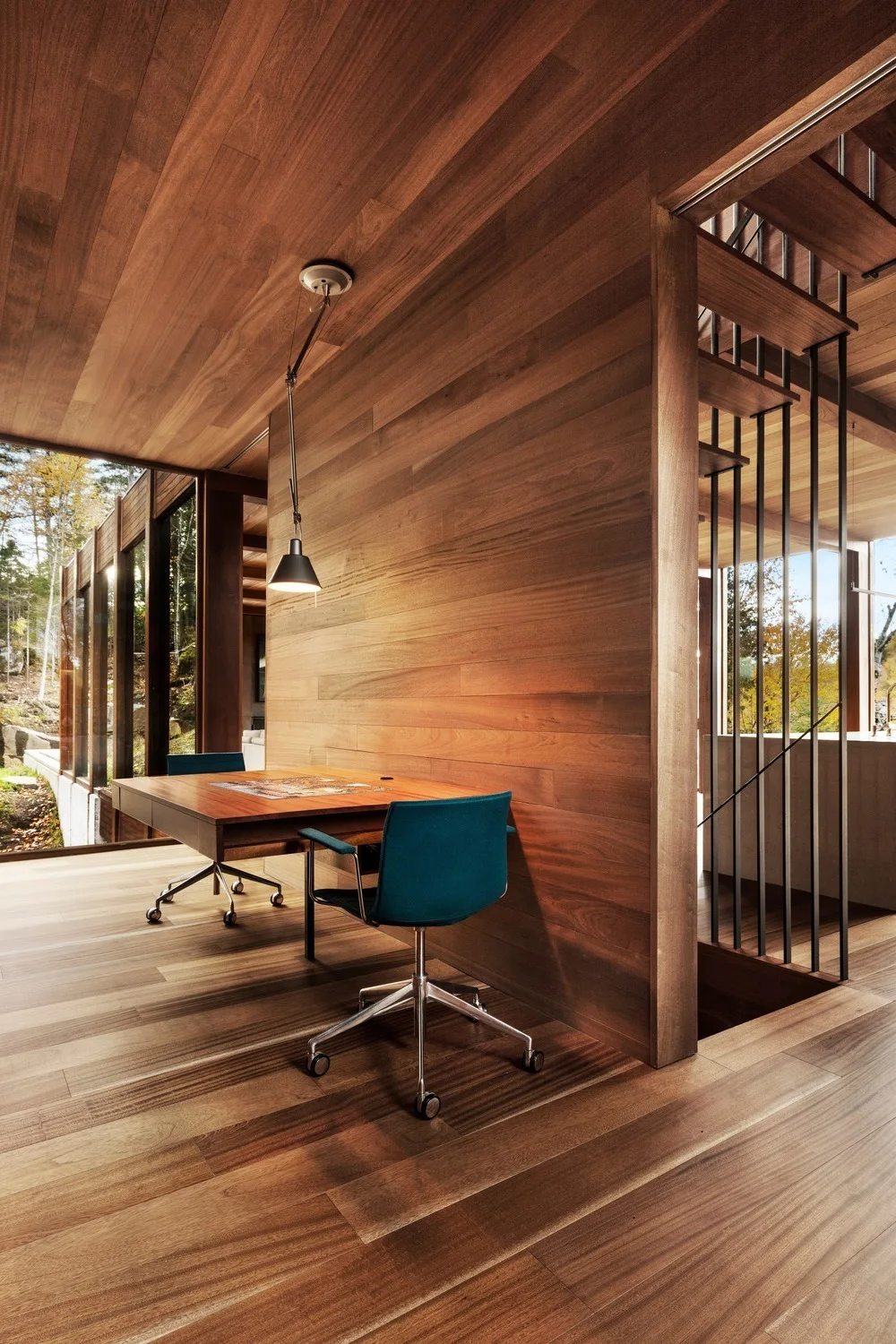 A modern wood-paneled office space features a warm wooden desk, teal upholstered chairs, and a black pendant light beside a staircase.