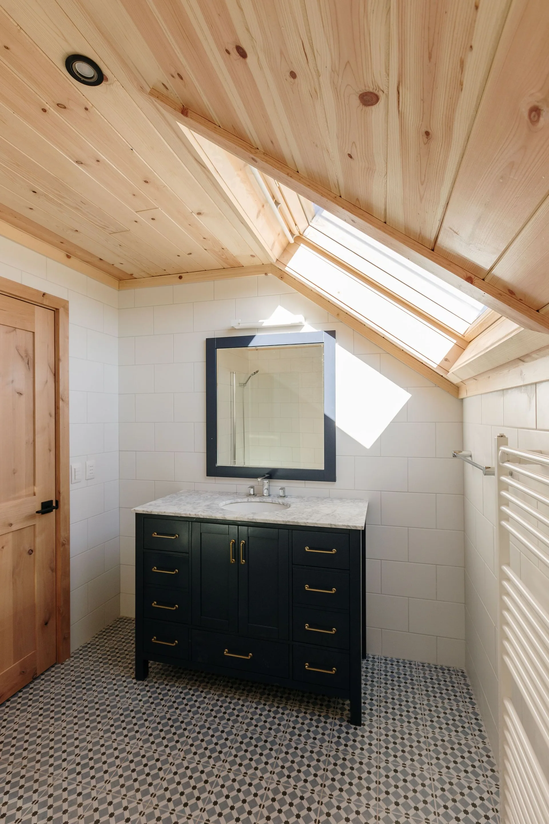 A modern attic bathroom featuring a dark blue vanity, white subway tile walls, patterned floor tiles, and a sloped wood plank ceiling with a skylight.