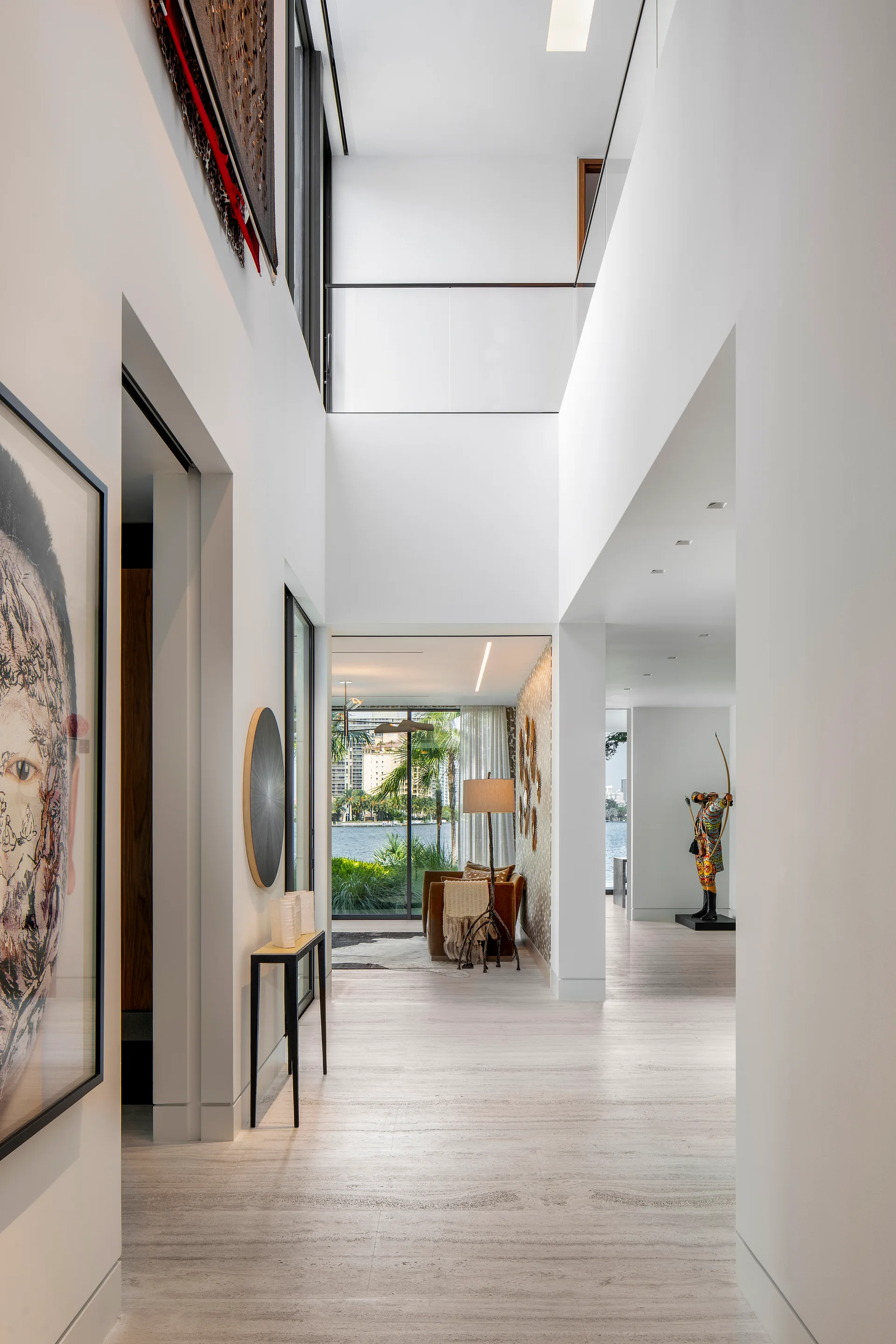 A bright double-height hallway features white walls, stone tile floors, a slim black console table, and a view leading to a waterfront seating area.