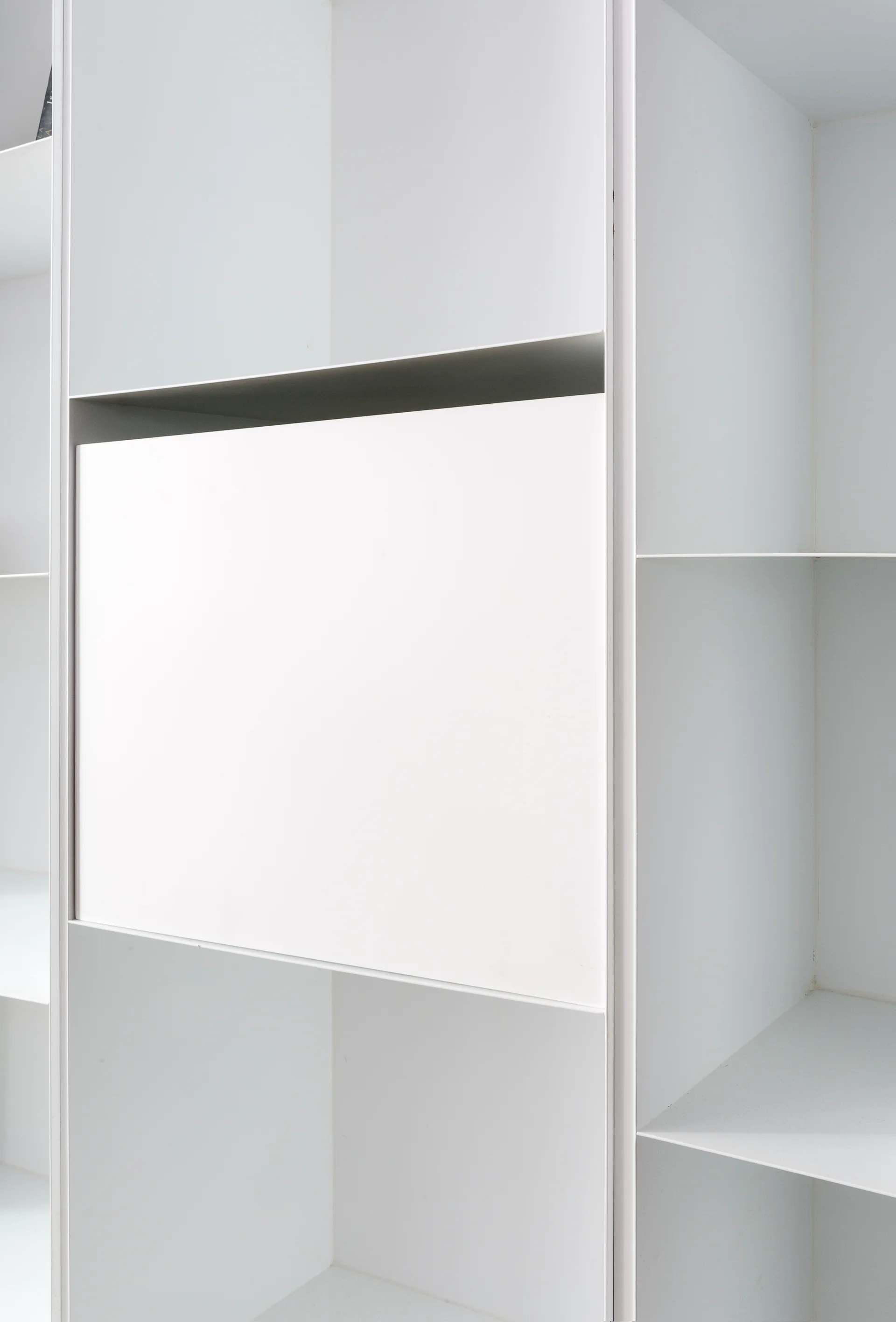 A minimalist matte white modular shelving unit featuring asymmetrical geometric open shelves and a single flush handleless cabinet panel.