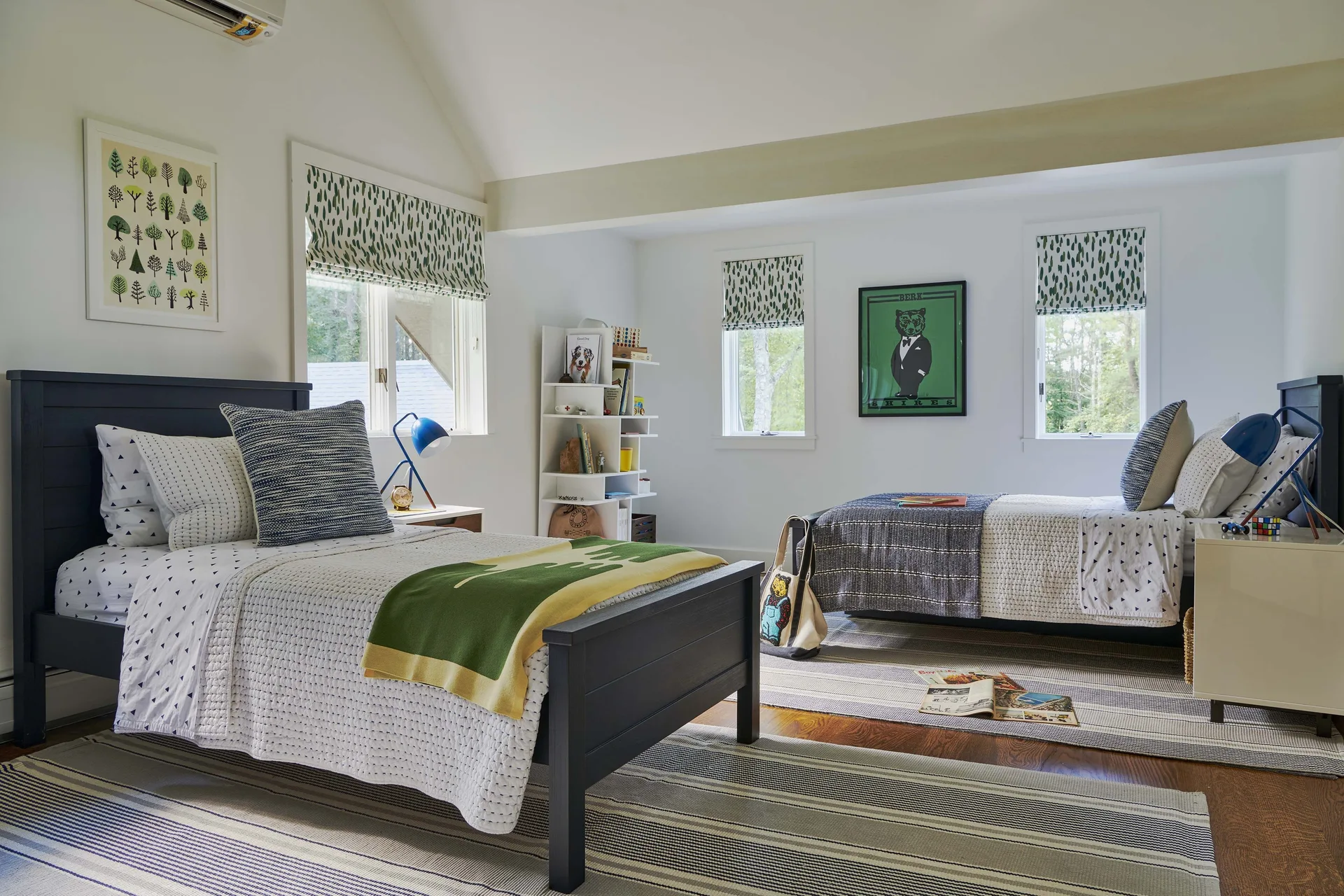 A shared children's bedroom featuring two dark wooden twin beds, striped rugs, white walls, and green patterned roman shades.