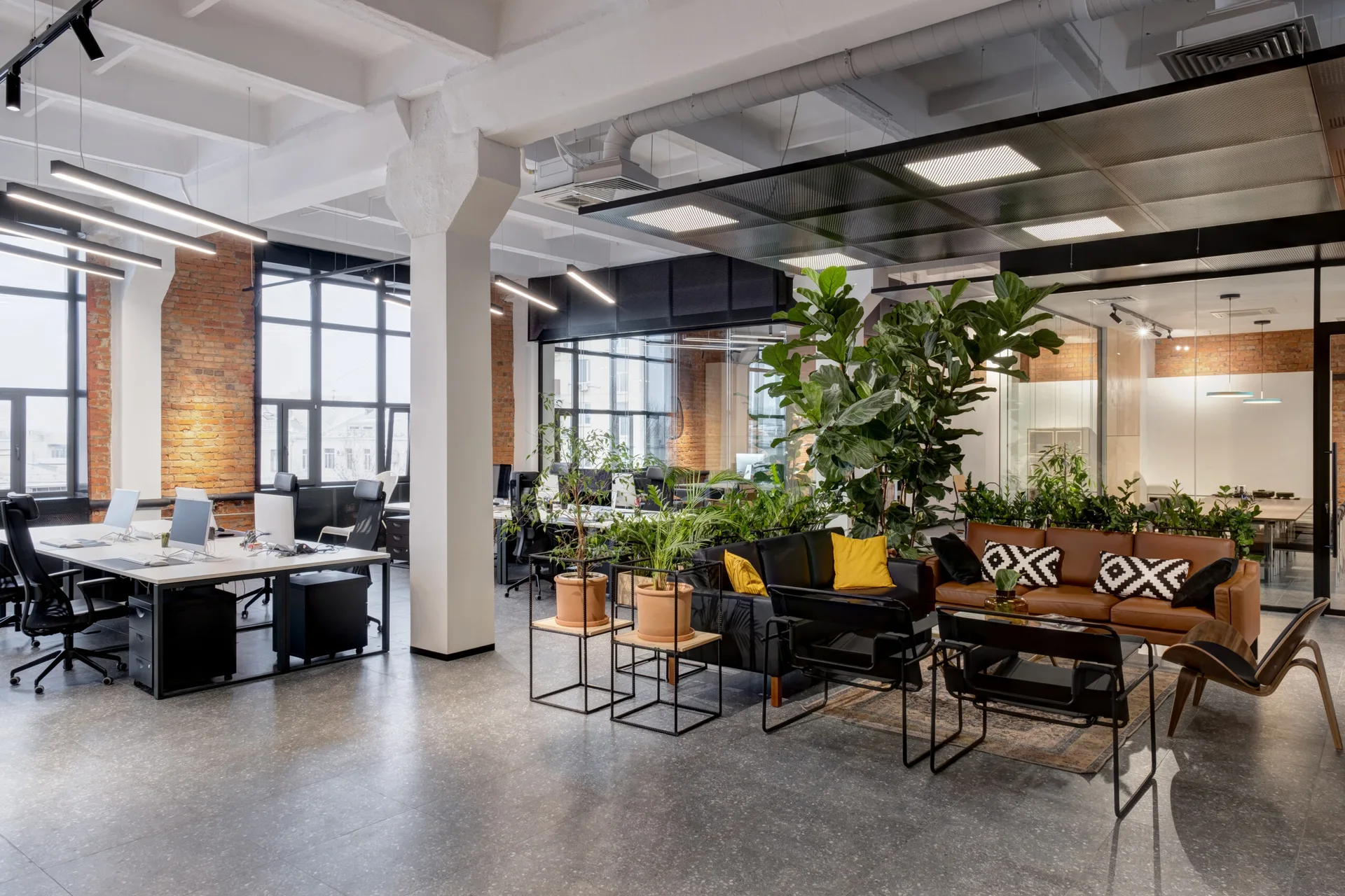 An open plan industrial office featuring white workstations, a comfortable lounge area with leather sofas, and numerous tall green houseplants.