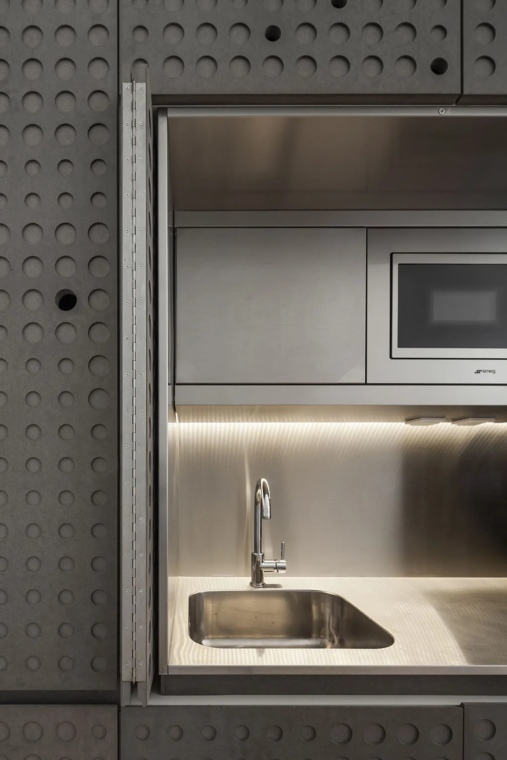 A compact stainless steel kitchenette with a small sink and integrated microwave, surrounded by textured dark grey perforated wall panels.