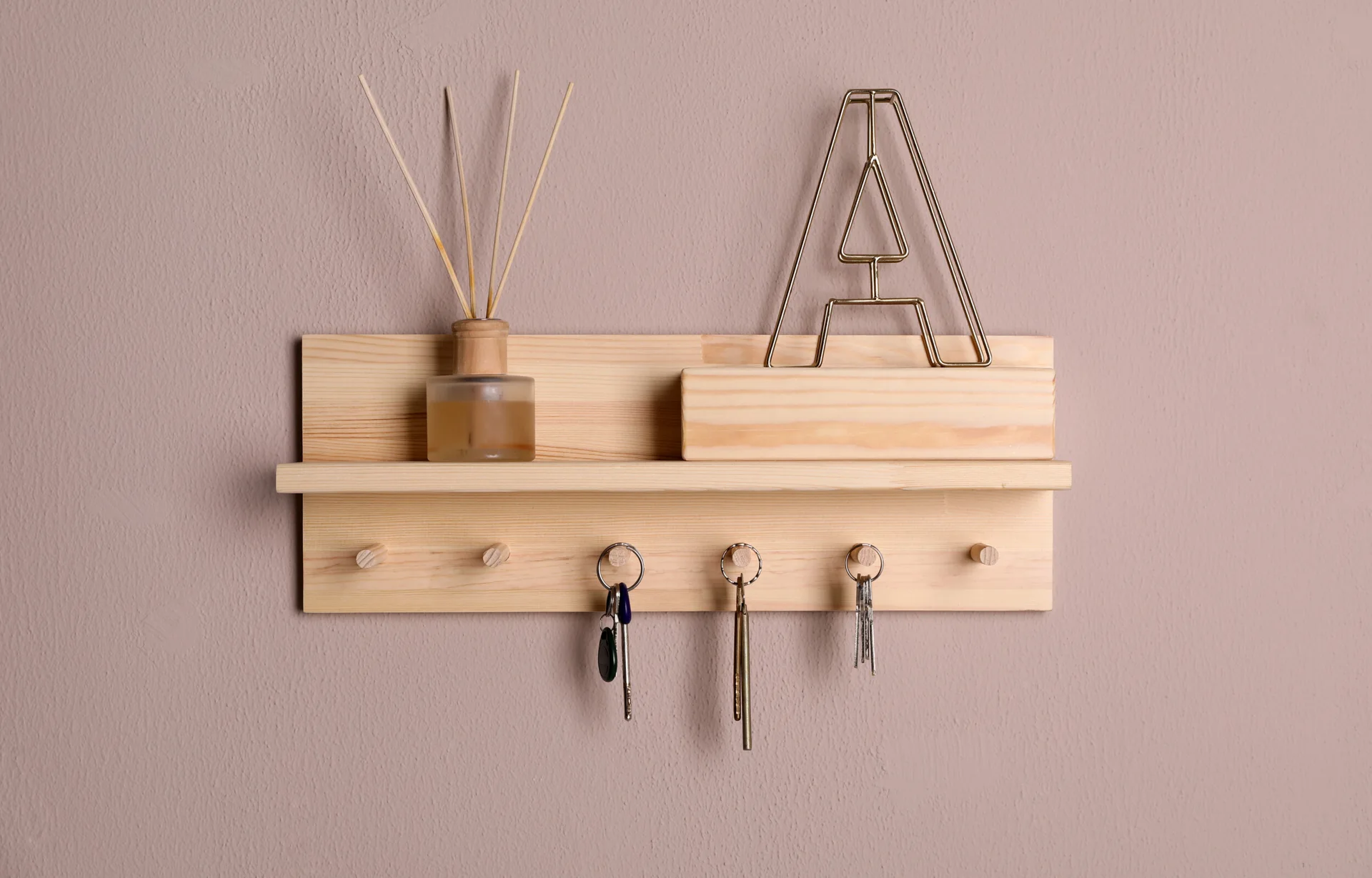 A light wood wall-mounted shelf featuring integrated key hooks, a reed diffuser, and a metal letter decoration against a matte mauve wall.