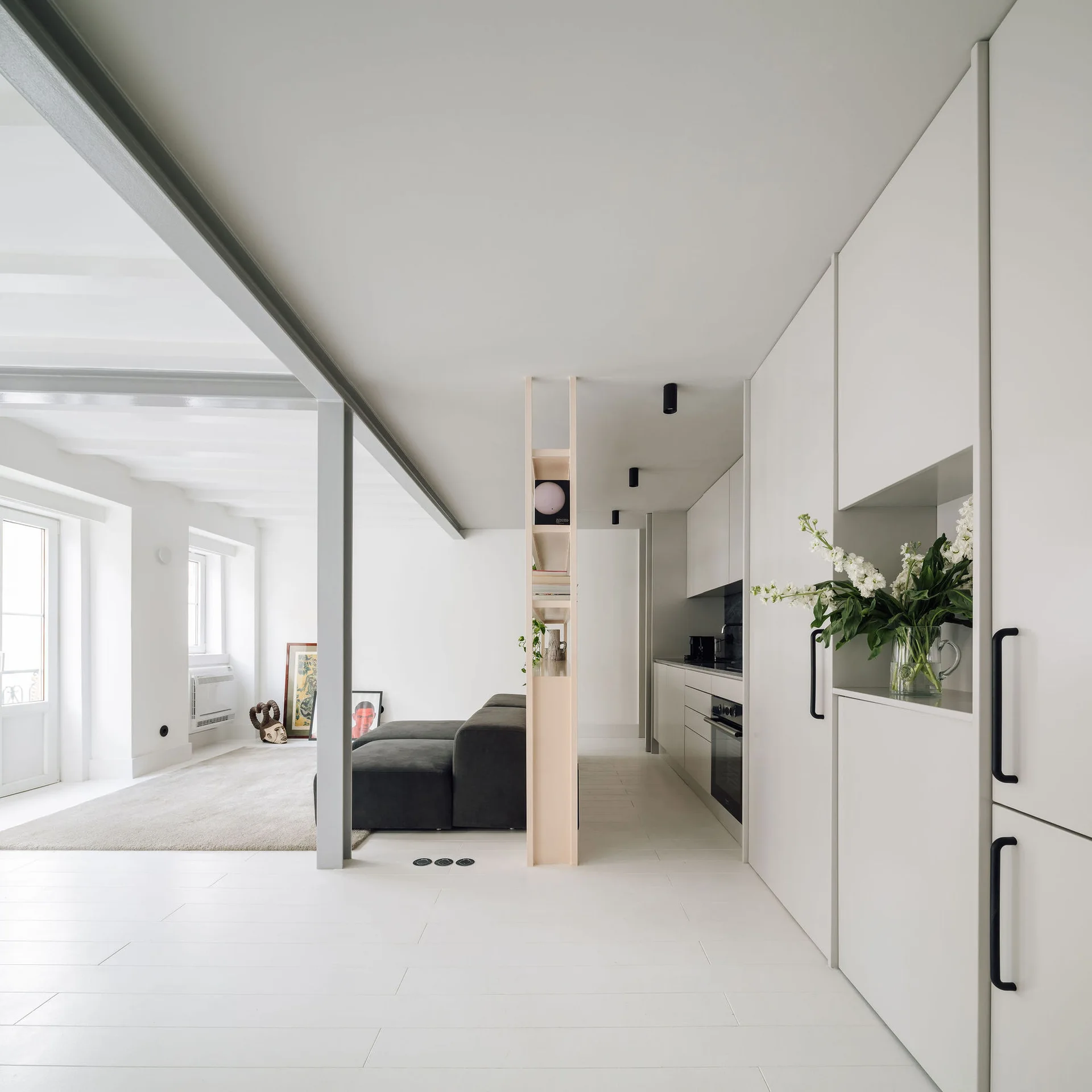 A minimalist open-plan kitchen and living room featuring white cabinetry with black handles, a dark grey sofa, and a slim wood divider shelf.