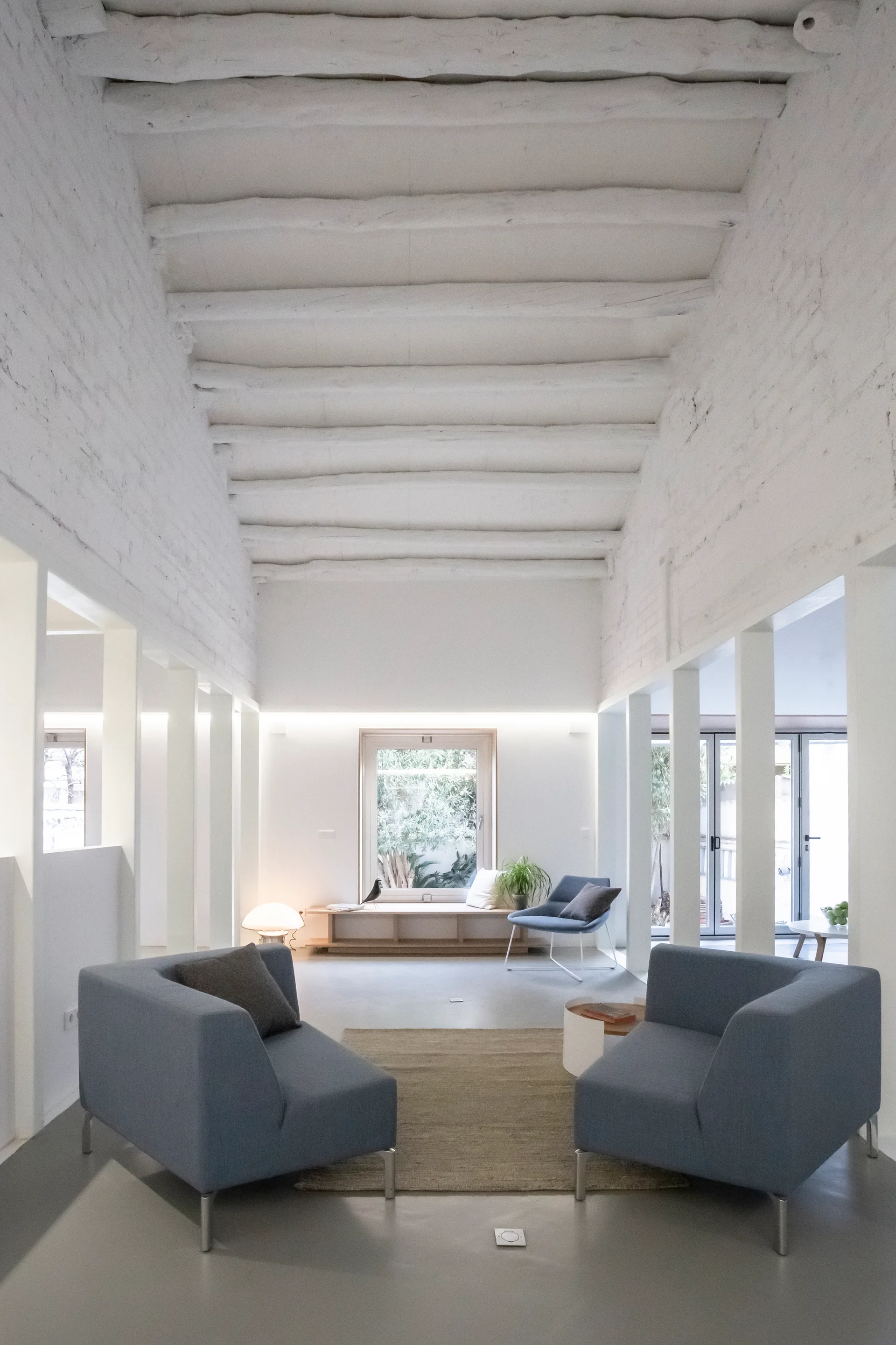 A minimalist white living room features two gray armchairs under a high ceiling with exposed beams and a row of vertical pillars.