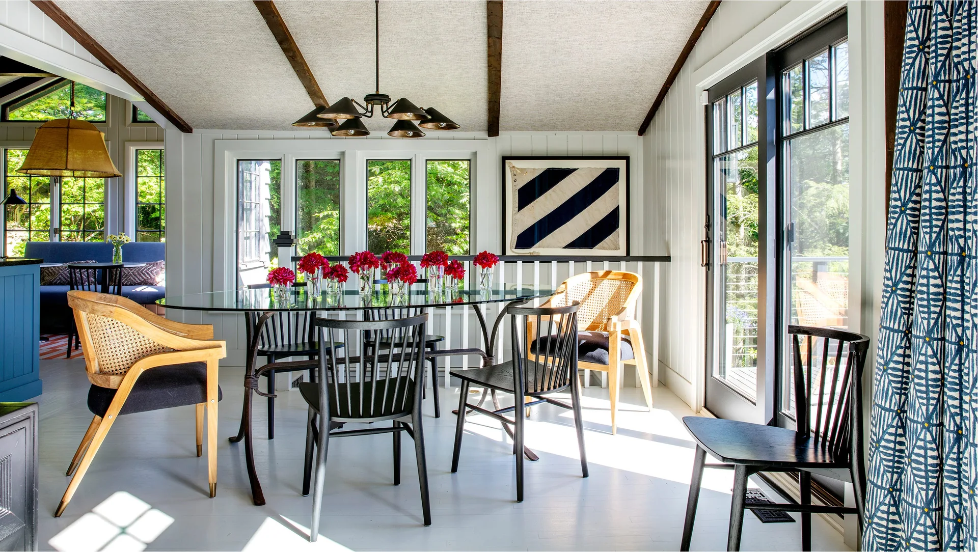 A light-filled dining room features a glass-topped table surrounded by black spindle and cane chairs beneath a vaulted ceiling with dark wood beams.
