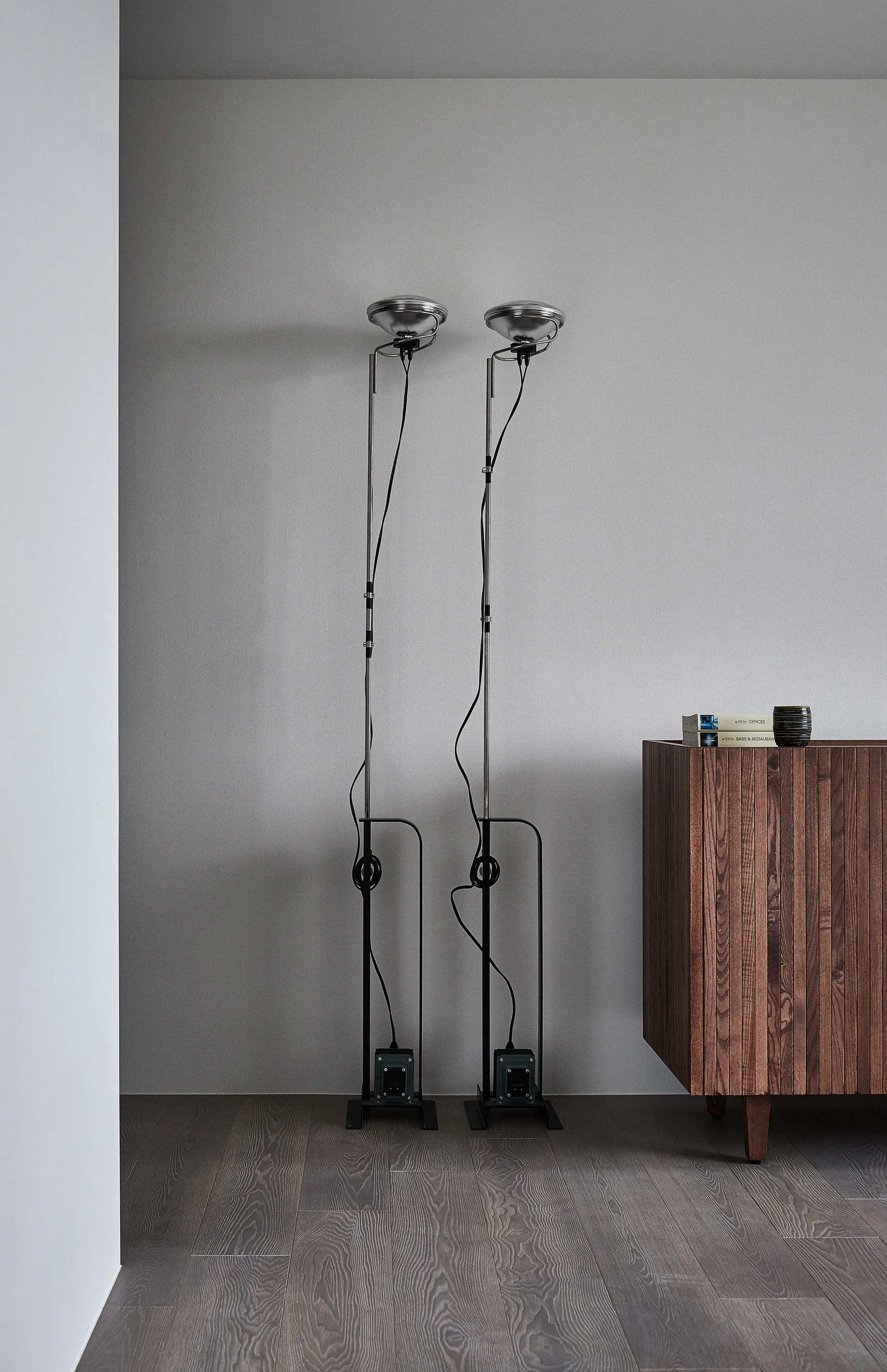 Two tall industrial floor lamps stand next to a slatted wooden cabinet against a plain grey wall on dark wood flooring.