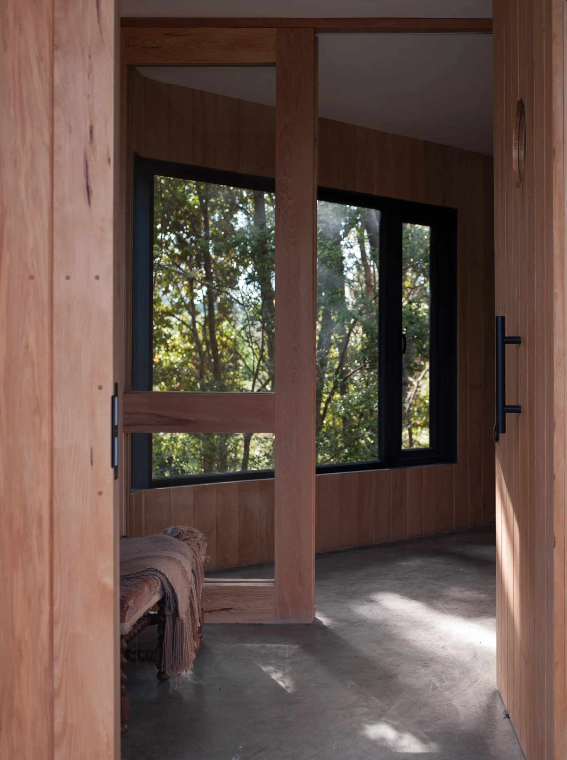 A concrete-floored entryway features walls clad in vertical wood paneling and a large window looking out at green forest trees.