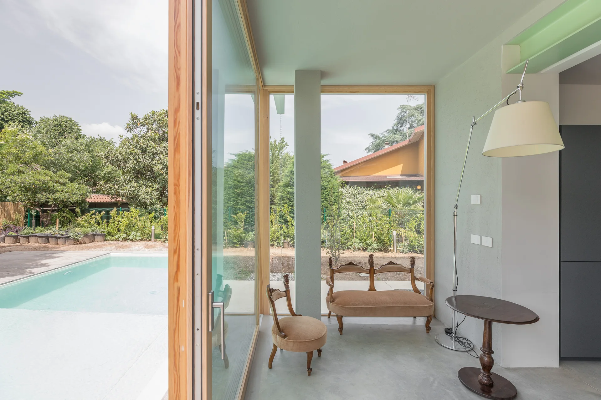 Antique upholstered chairs sit on a grey concrete floor next to floor-to-ceiling glass doors overlooking an outdoor swimming pool.