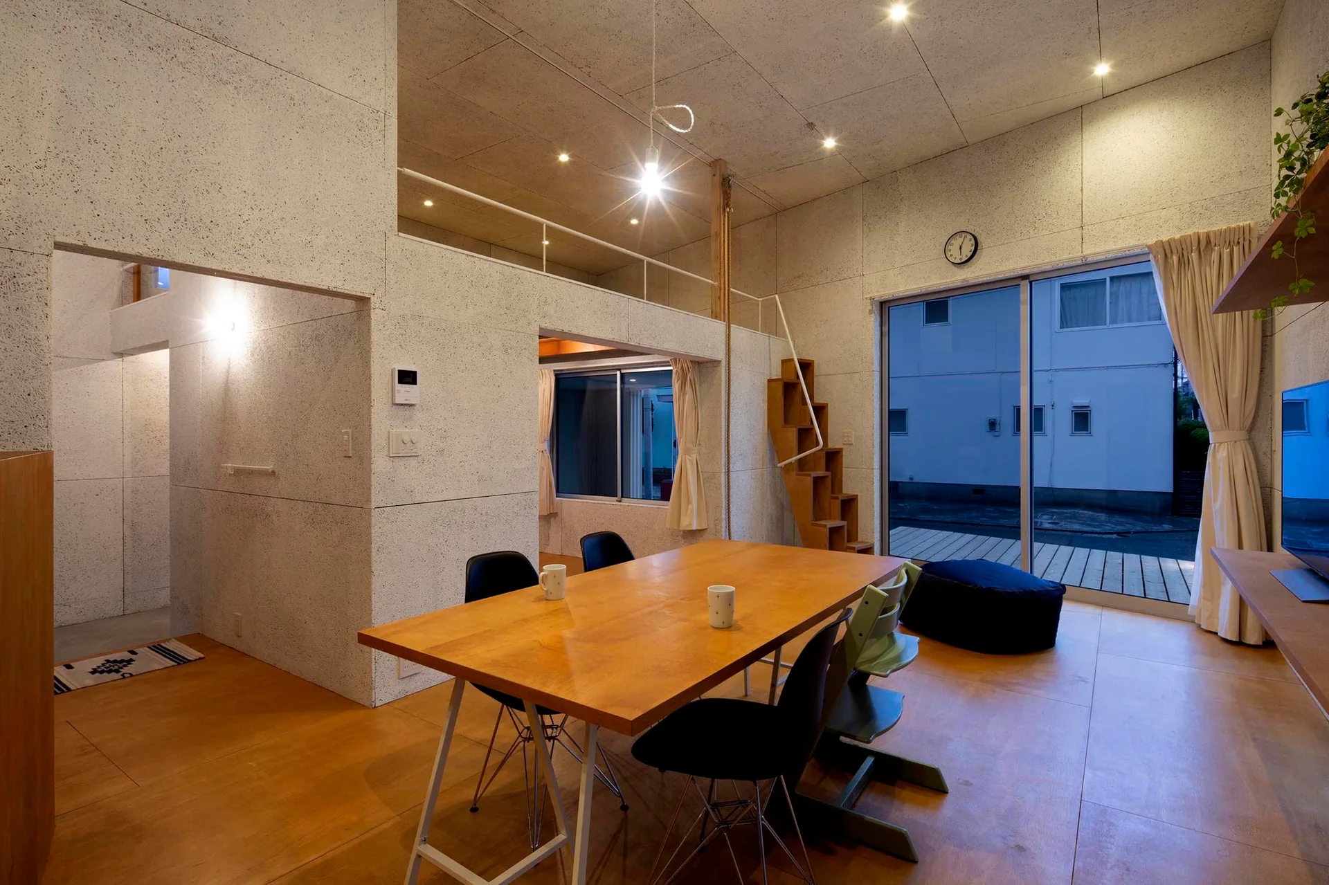 A dining room features a wooden table with black chairs, textured light grey walls, and a wooden open-riser staircase leading to a loft.