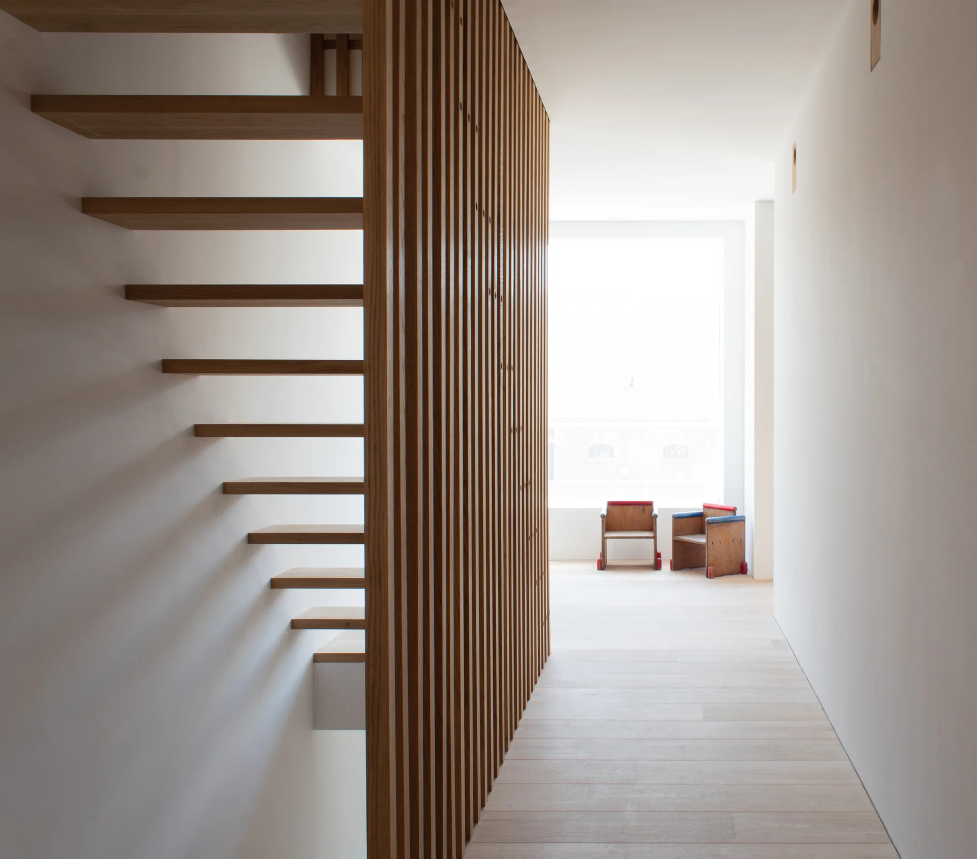 A minimalist hallway features a floating wood staircase next to a vertical slatted partition wall with a bright window at the end.