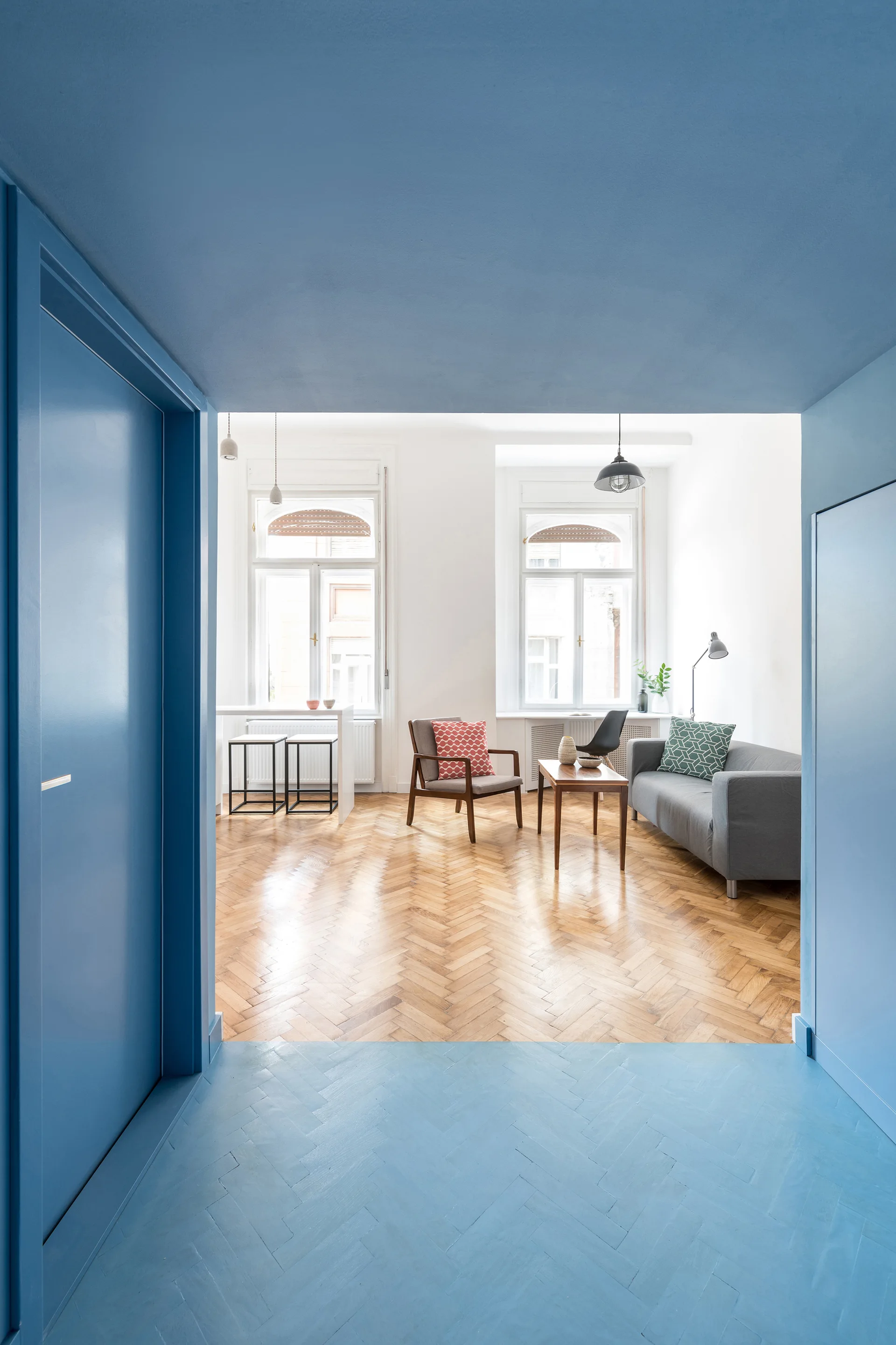 A view from a blue-painted entryway looking into a bright living room with herringbone parquet floors, a grey sofa, and mid-century furniture.