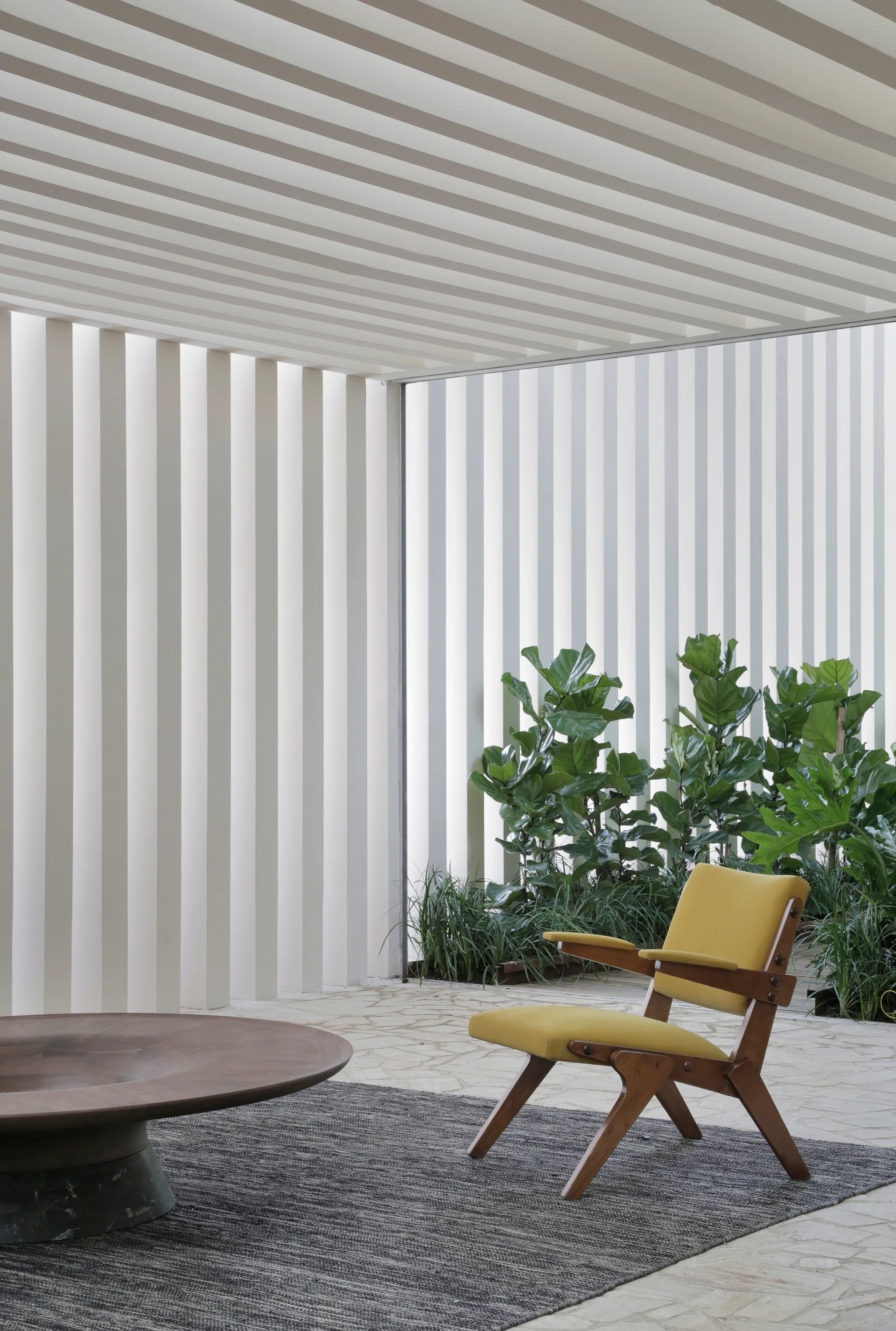 A yellow mid-century armchair and a round wooden coffee table sit on a grey rug against a backdrop of white vertical slats and green plants.