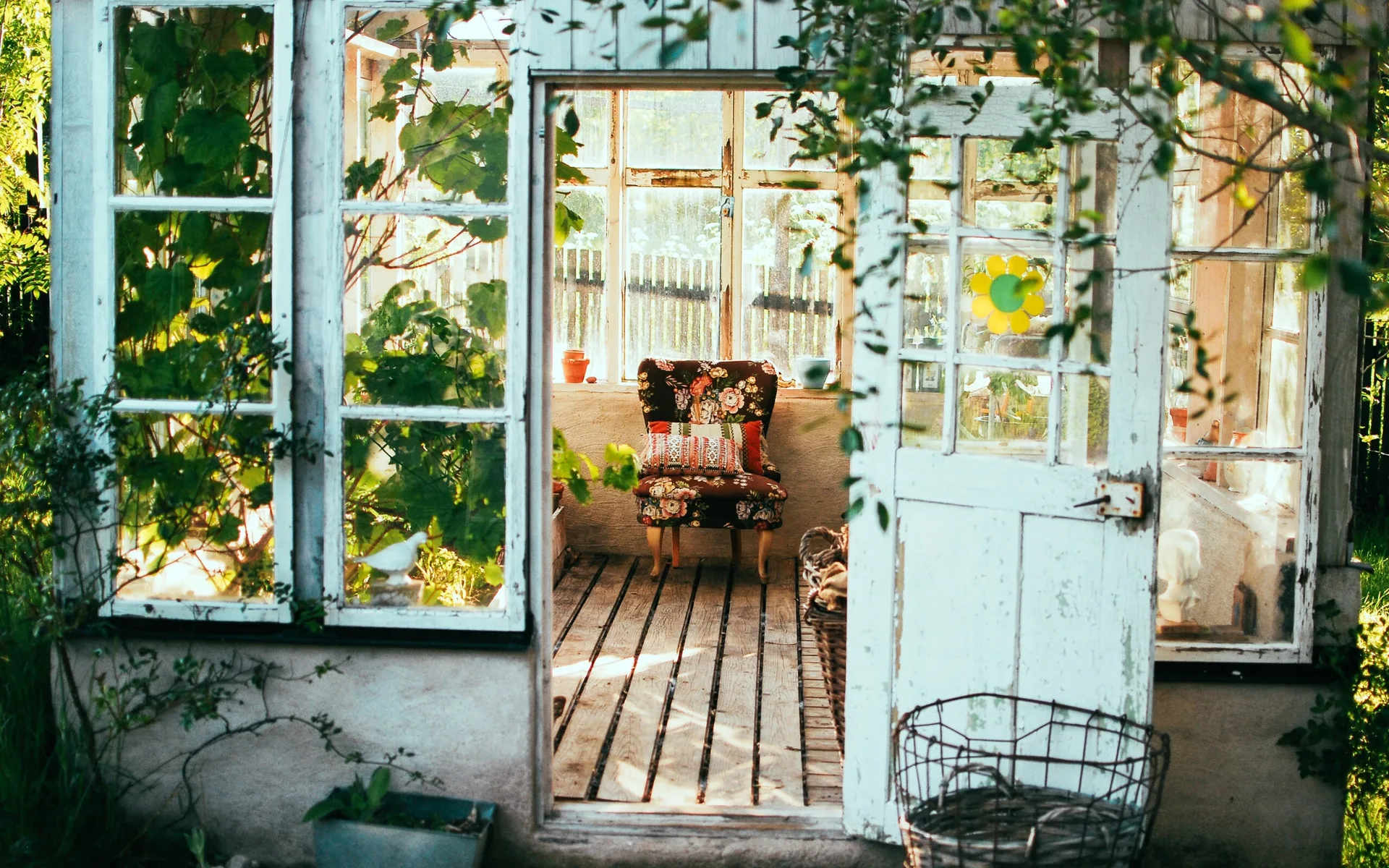 An open wooden door leads into a rustic greenhouse where a floral patterned armchair sits on wooden floorboards surrounded by glass walls.