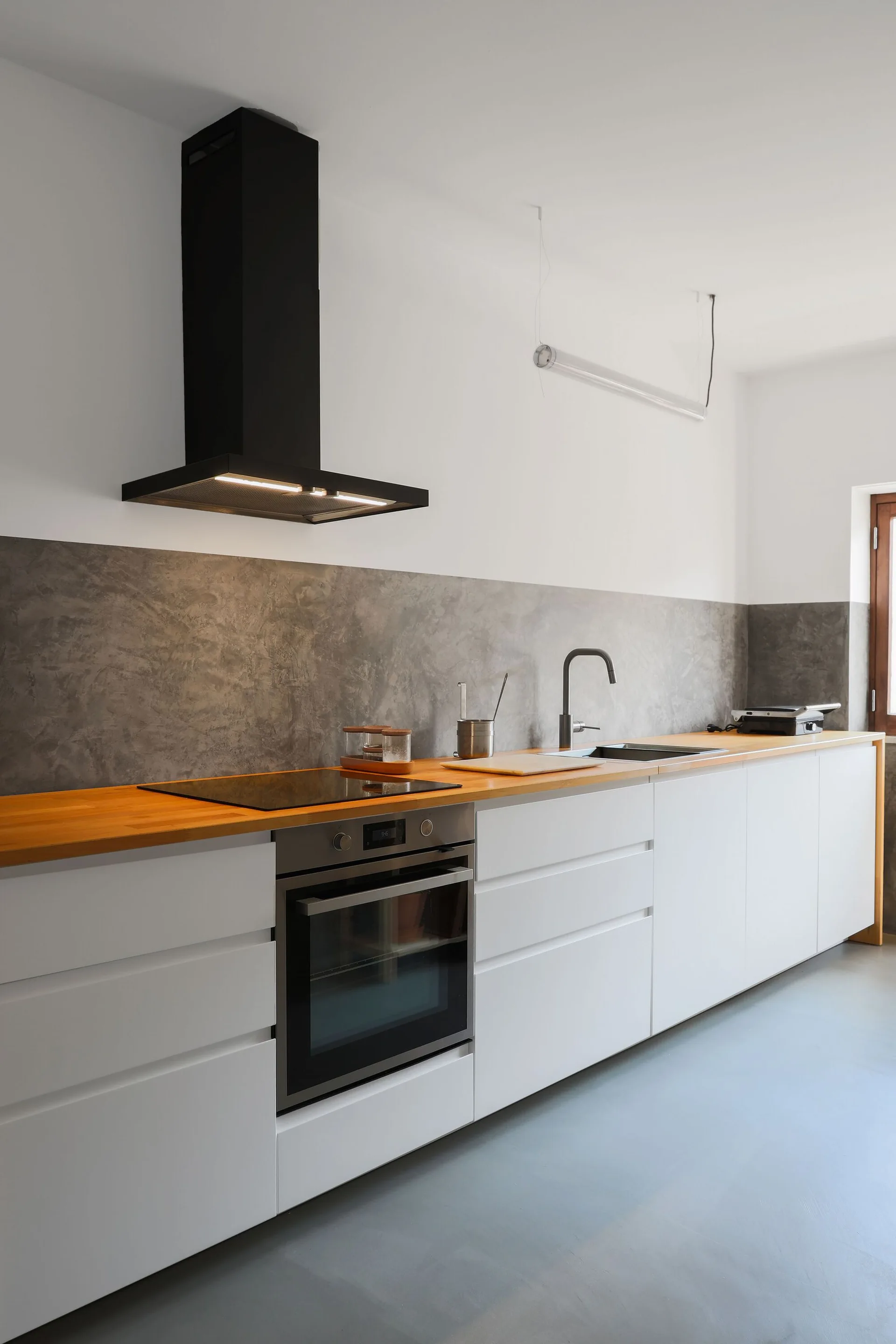 A modern kitchen features white flat-front lower cabinets, a warm wood countertop, a textured grey backsplash, and a black range hood.