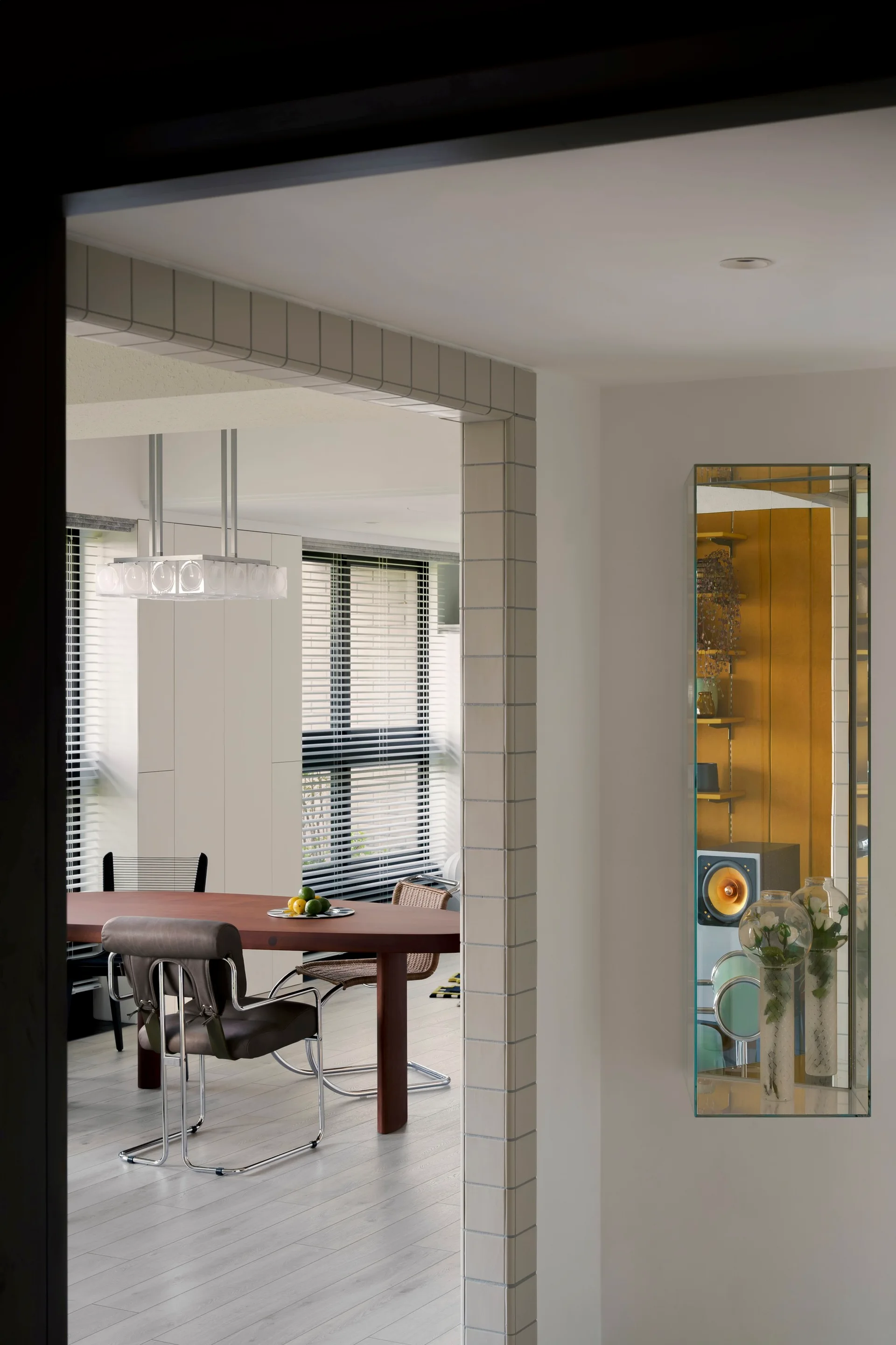 A modern dining room viewed through a square-tiled archway features an oval wood table, chrome cantilever chairs, and a narrow wall mirror.