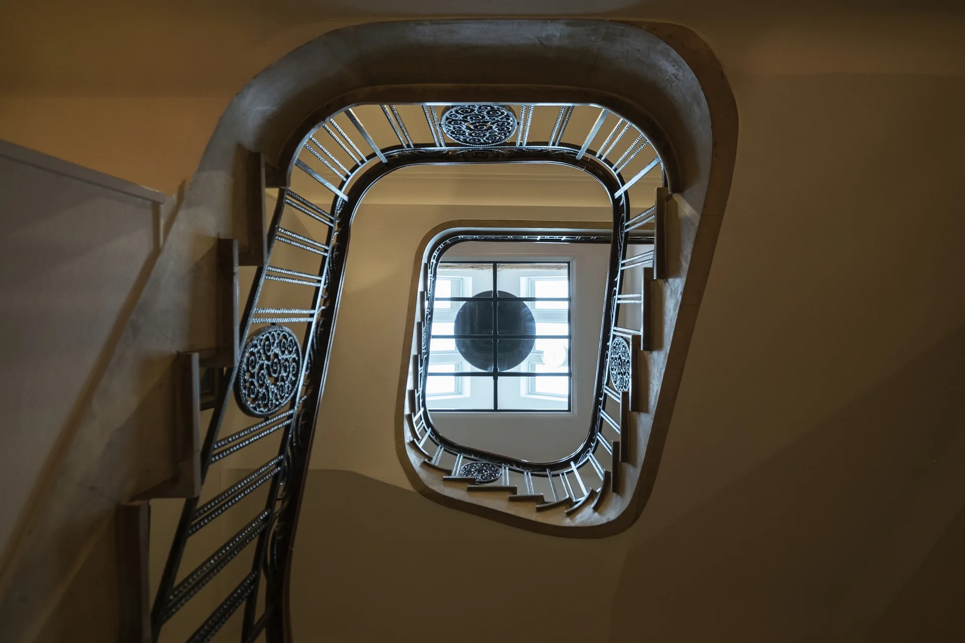A low-angle view looking up a curved spiral staircase with ornate black metal railings toward a bright central skylight.