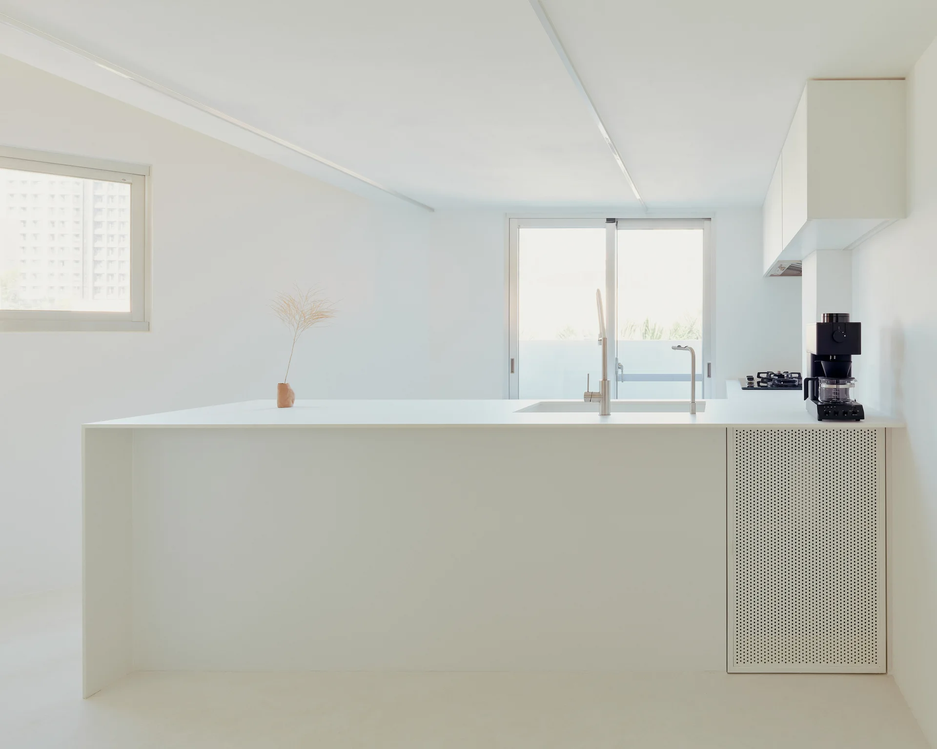 A minimalist white kitchen features a central island with an integrated sink, faucet, and a perforated white metal cabinet door on one side.