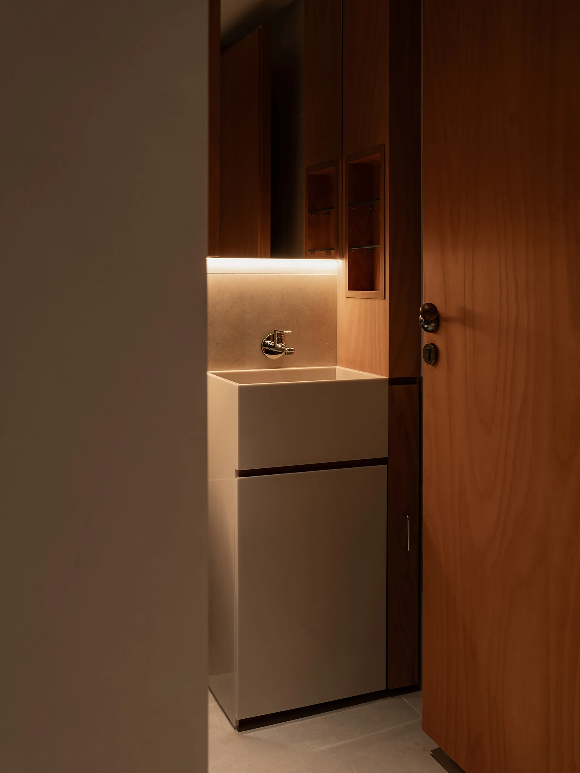 A minimalist powder room features a white block sink integrated into warm wood cabinetry, with a wall-mounted faucet and soft linear lighting.