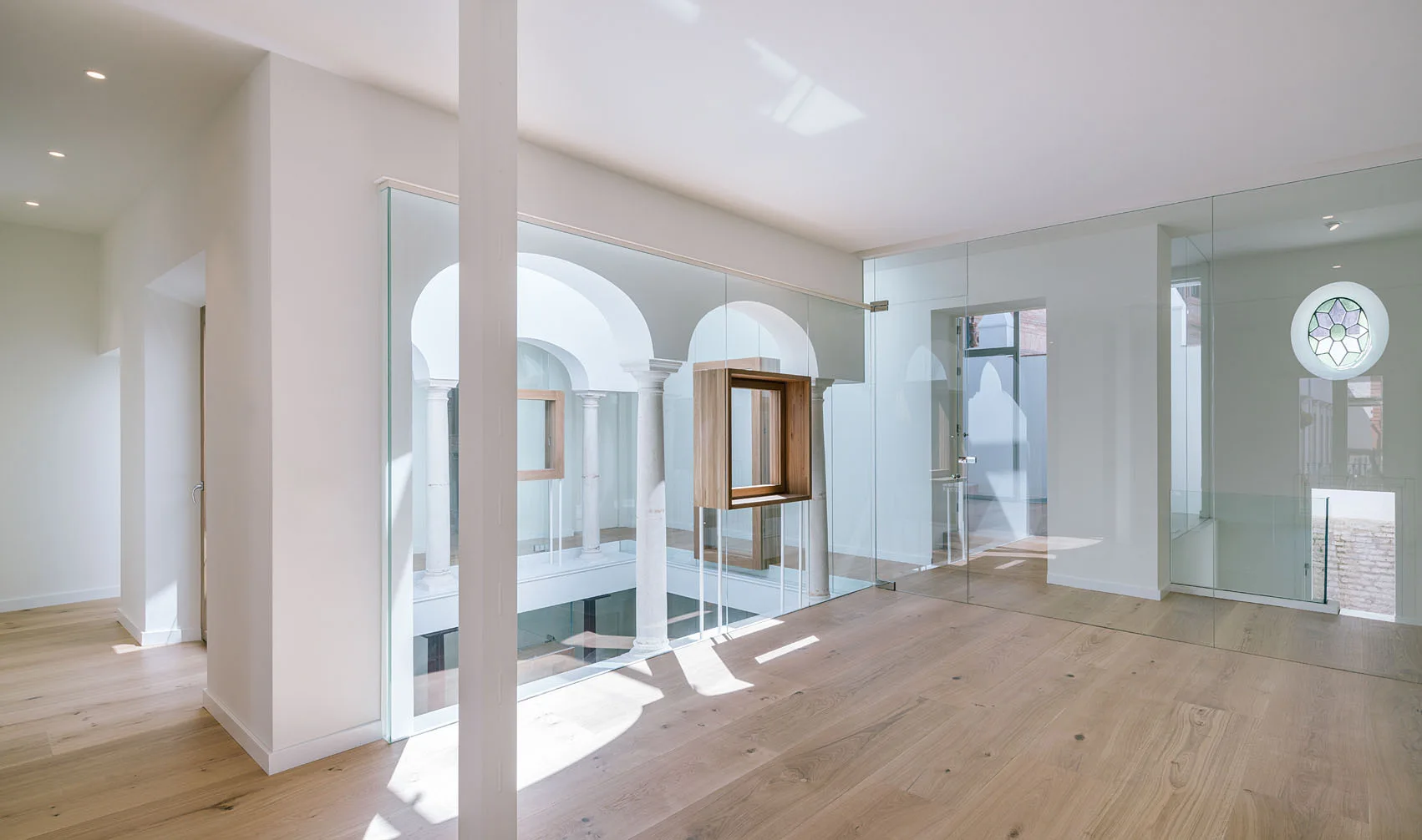 A bright minimalist landing features light wood floors, white walls, and frameless glass partitions overlooking historic stone arches and columns.