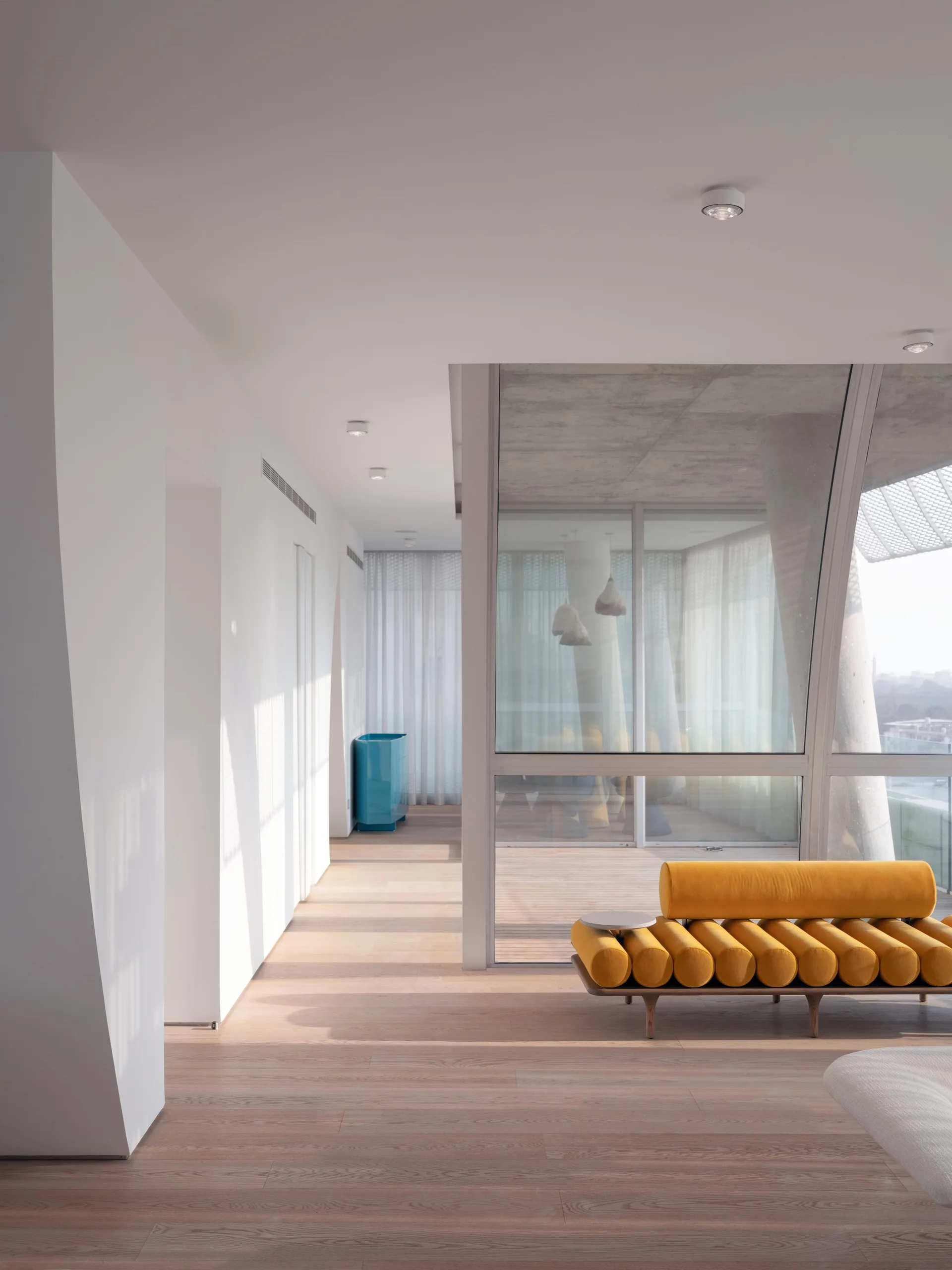 A minimalist living room features a yellow bolster cushion sofa next to large glass walls, with light wood floors and white walls.