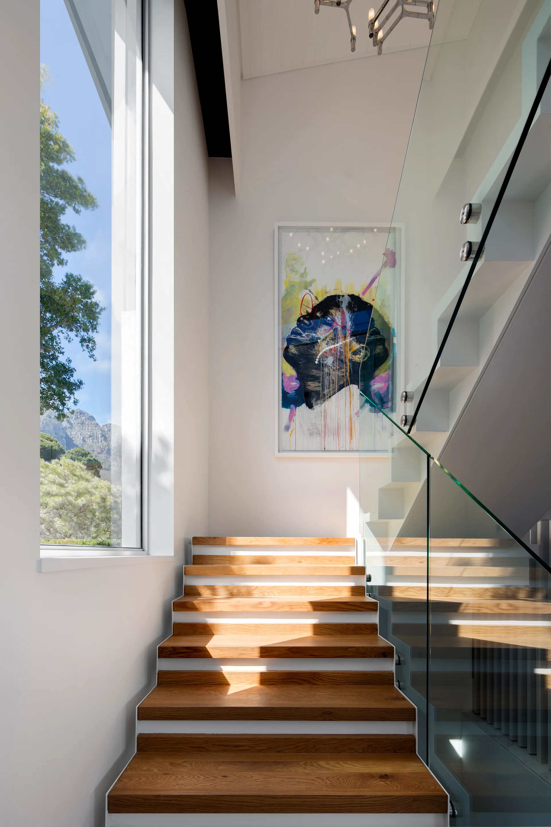 A modern staircase features warm wood treads, white risers, and a clear glass railing under a tall window showing mountain views.