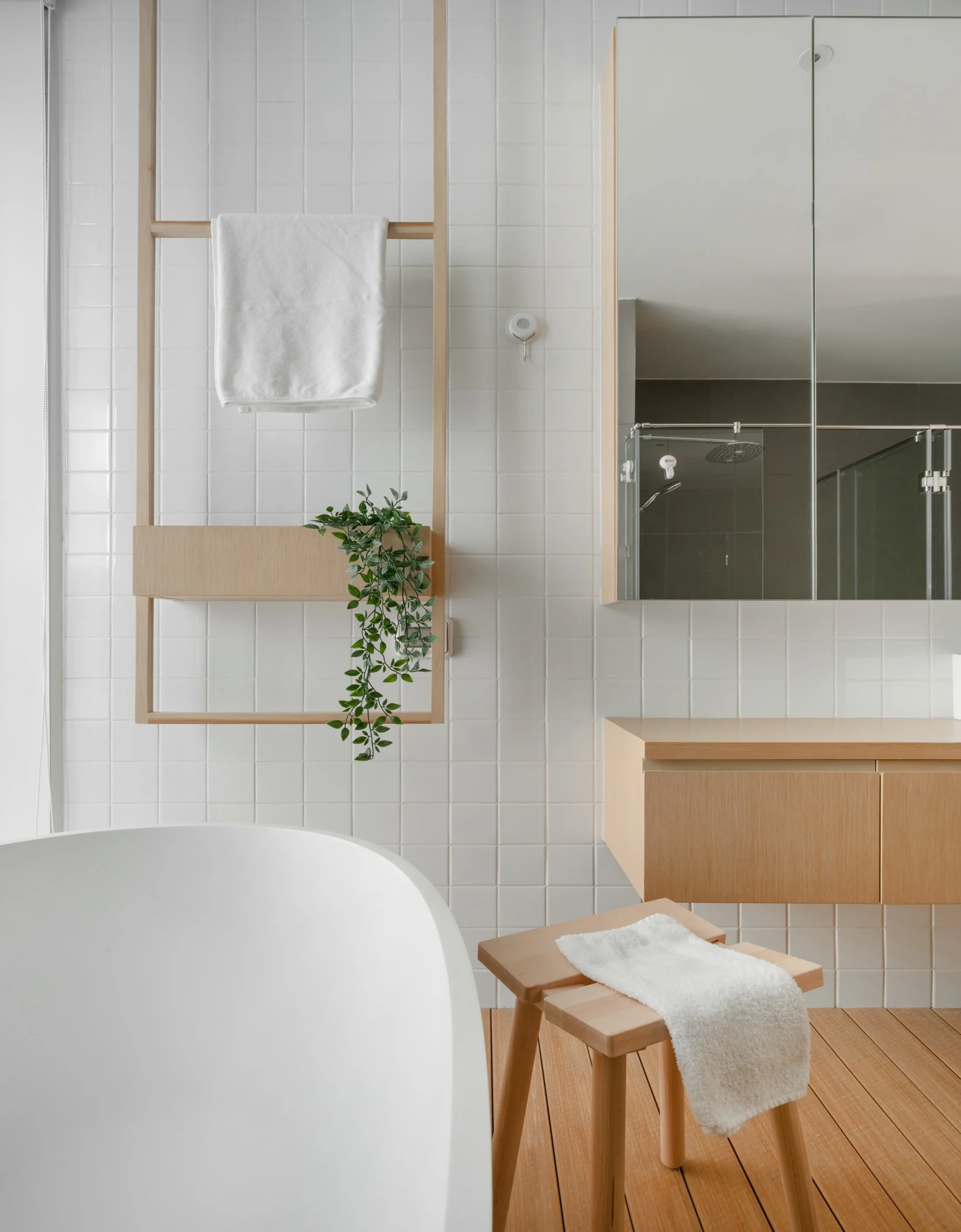 A minimalist bathroom featuring a white freestanding bathtub, square grid wall tiles, a light wood floating vanity, and a matching wooden floor.