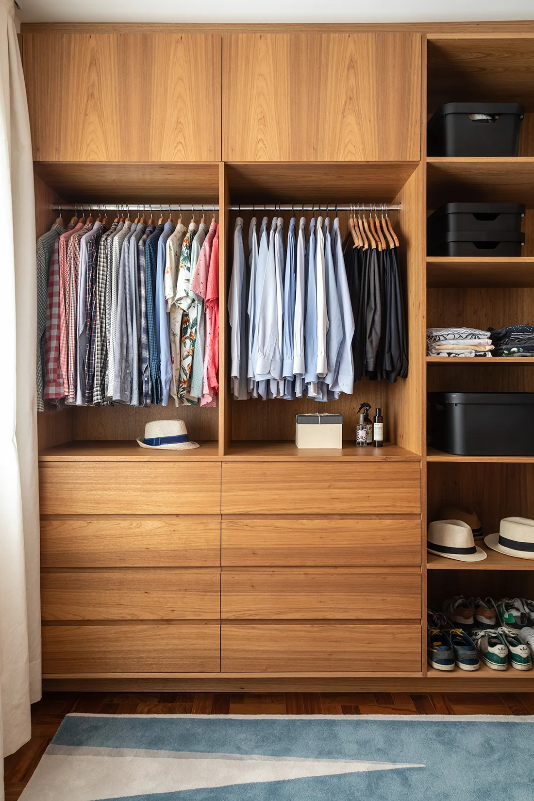 A custom built-in wood wardrobe closet features organized hanging shirts, flat-front drawers, and open shelving holding storage boxes above a blue rug.