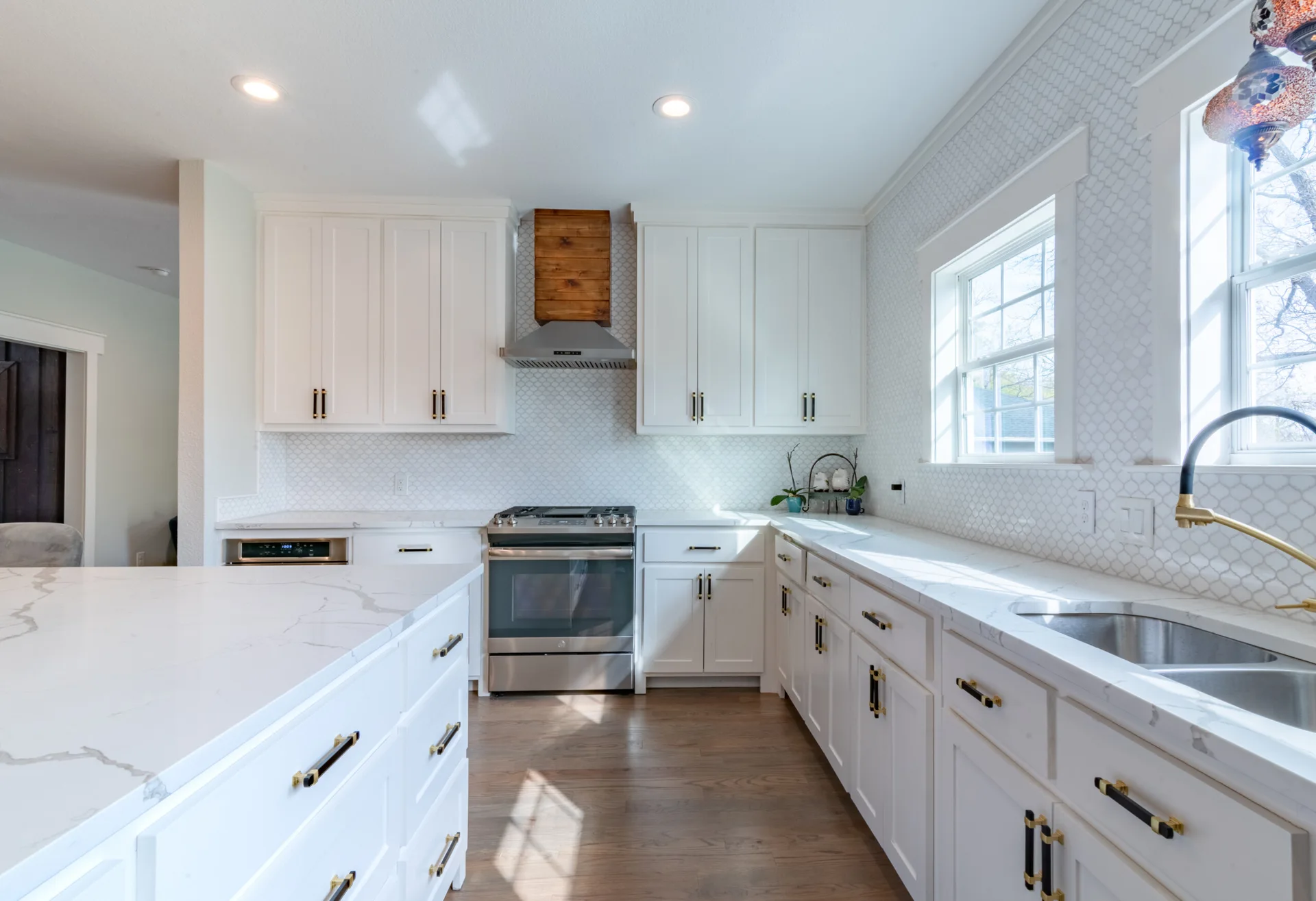 A bright kitchen featuring white shaker cabinets, an arabesque tile backsplash, a wood-accented range hood, and a marble-look island.