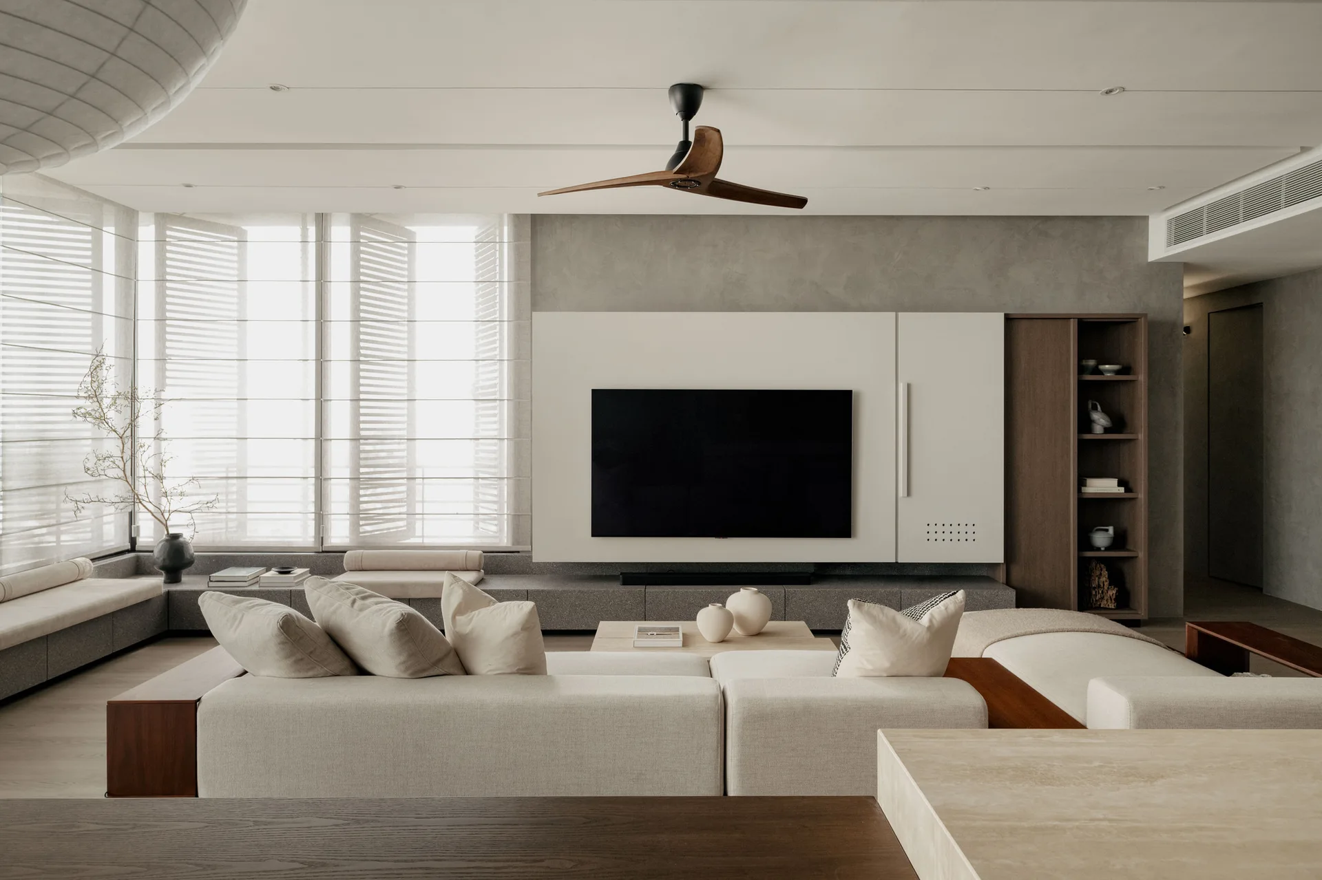 A minimalist living room featuring a low-profile cream sofa, a built-in white and wood media unit, and a long grey bench along a window with blinds.