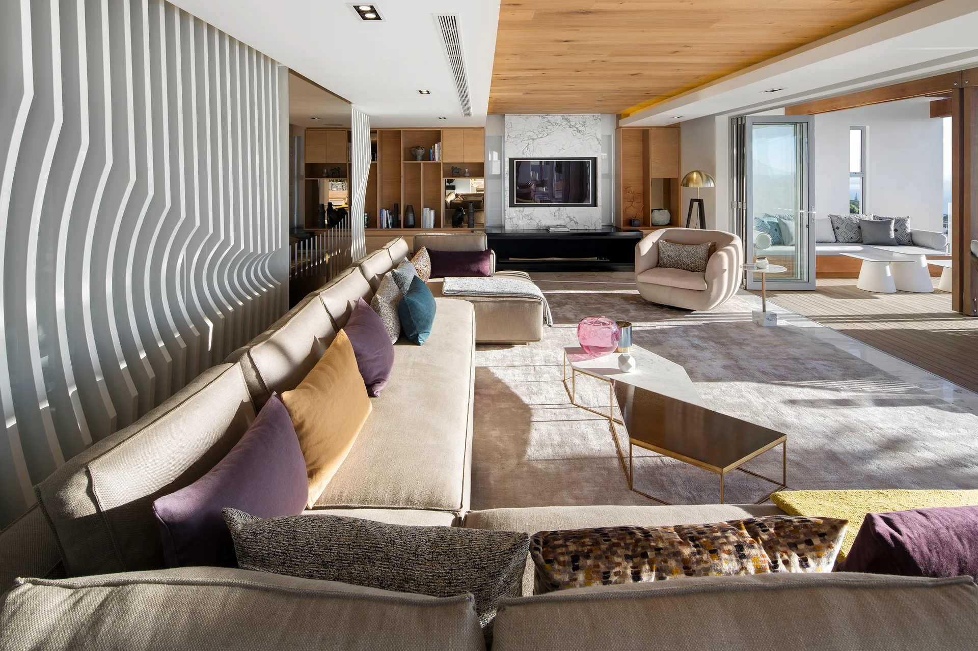 A modern living room features a tan sectional sofa, geometric coffee tables, a sculptural white wave wall, and a wood plank ceiling.