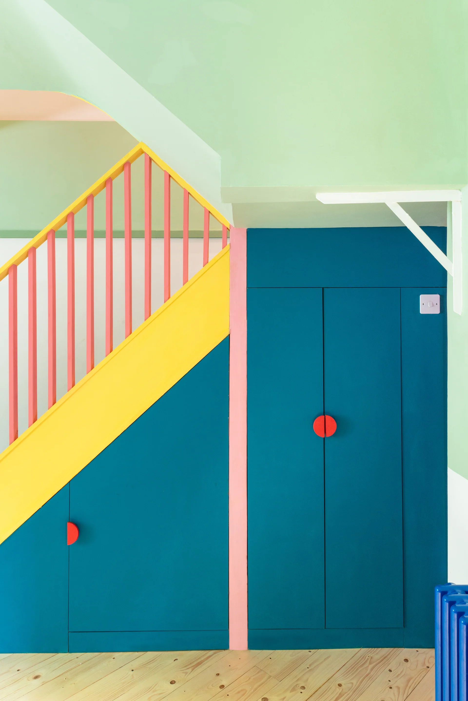 A color-blocked staircase with yellow trim and pink balusters features dark blue under-stair cabinets with circular red handles.