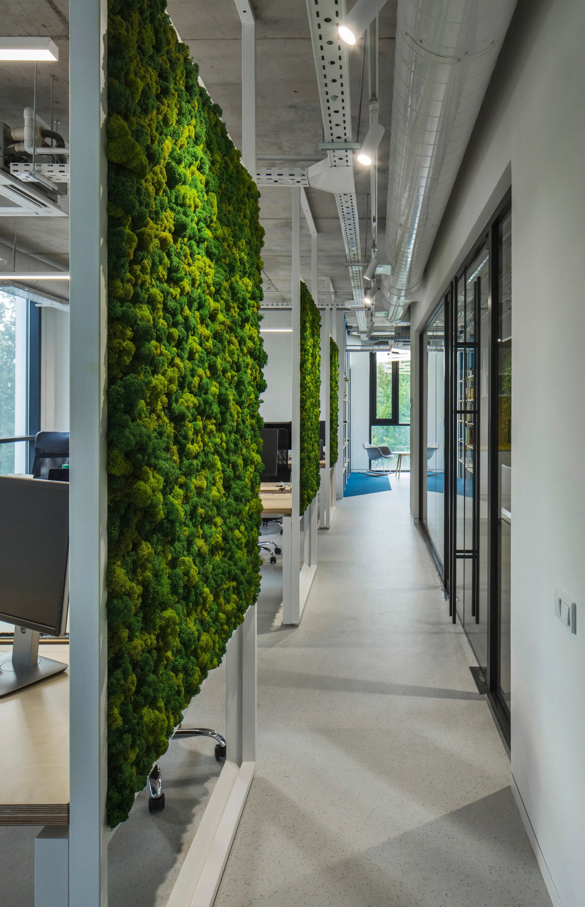 An open-plan office features green moss wall partitions separating workstations, with an exposed concrete ceiling, ductwork, and track lighting.