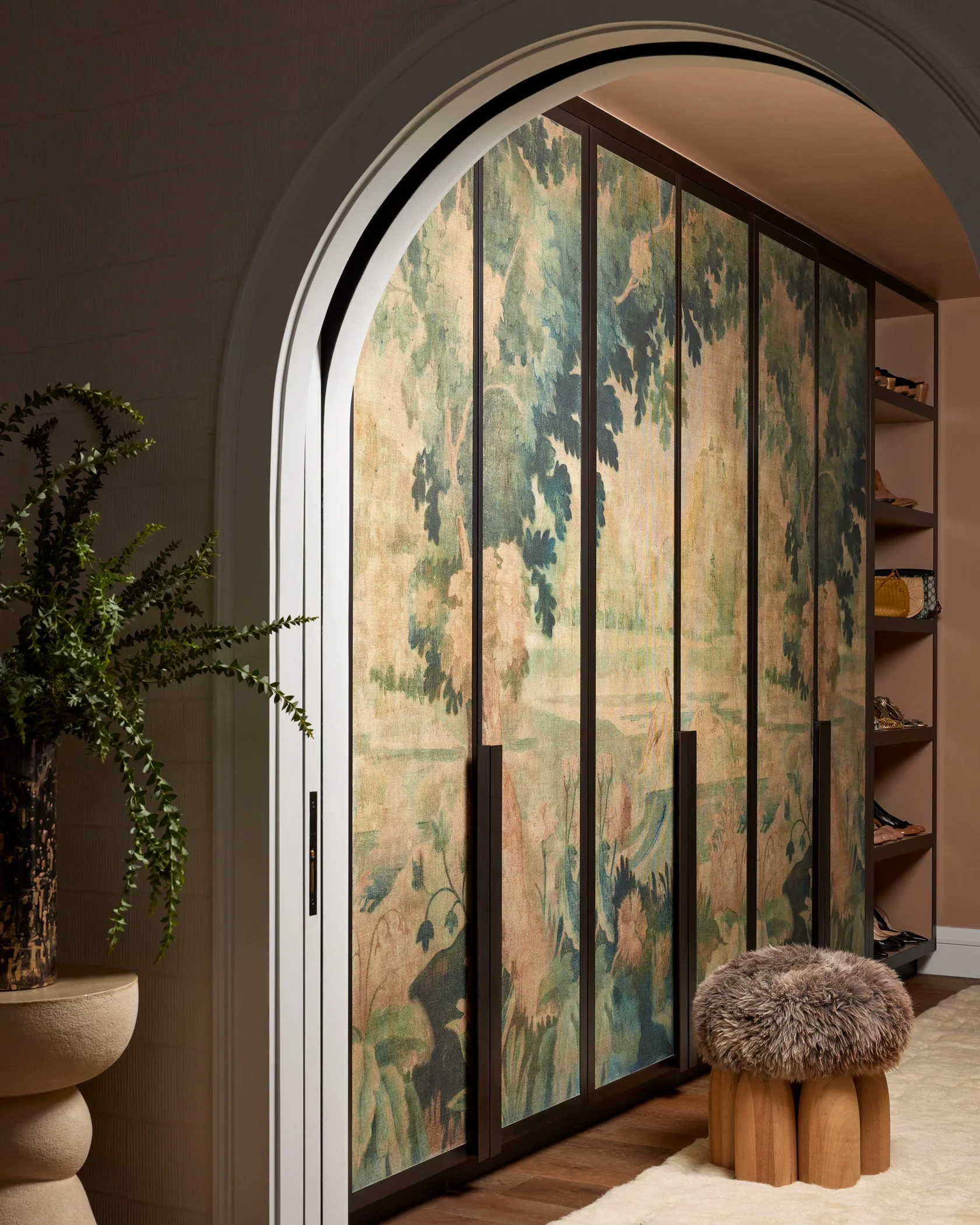 An arched opening reveals custom wardrobe doors featuring a green forest scenic mural alongside a furry wooden stool and open shelving.