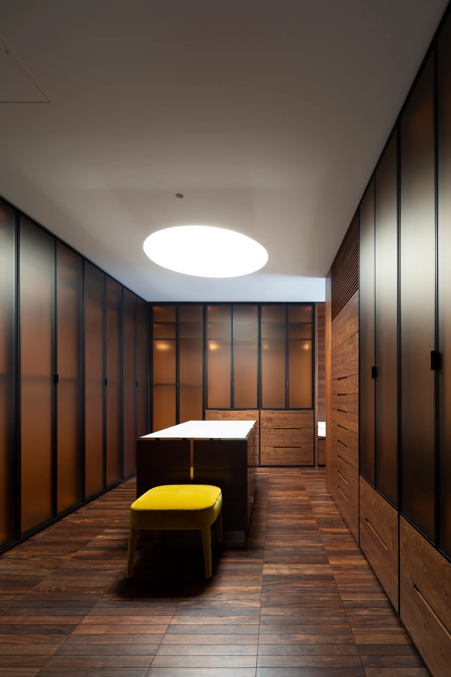 A modern dressing room featuring dark glass-fronted wardrobes, a central island with a white top, a yellow stool, and a circular ceiling light.
