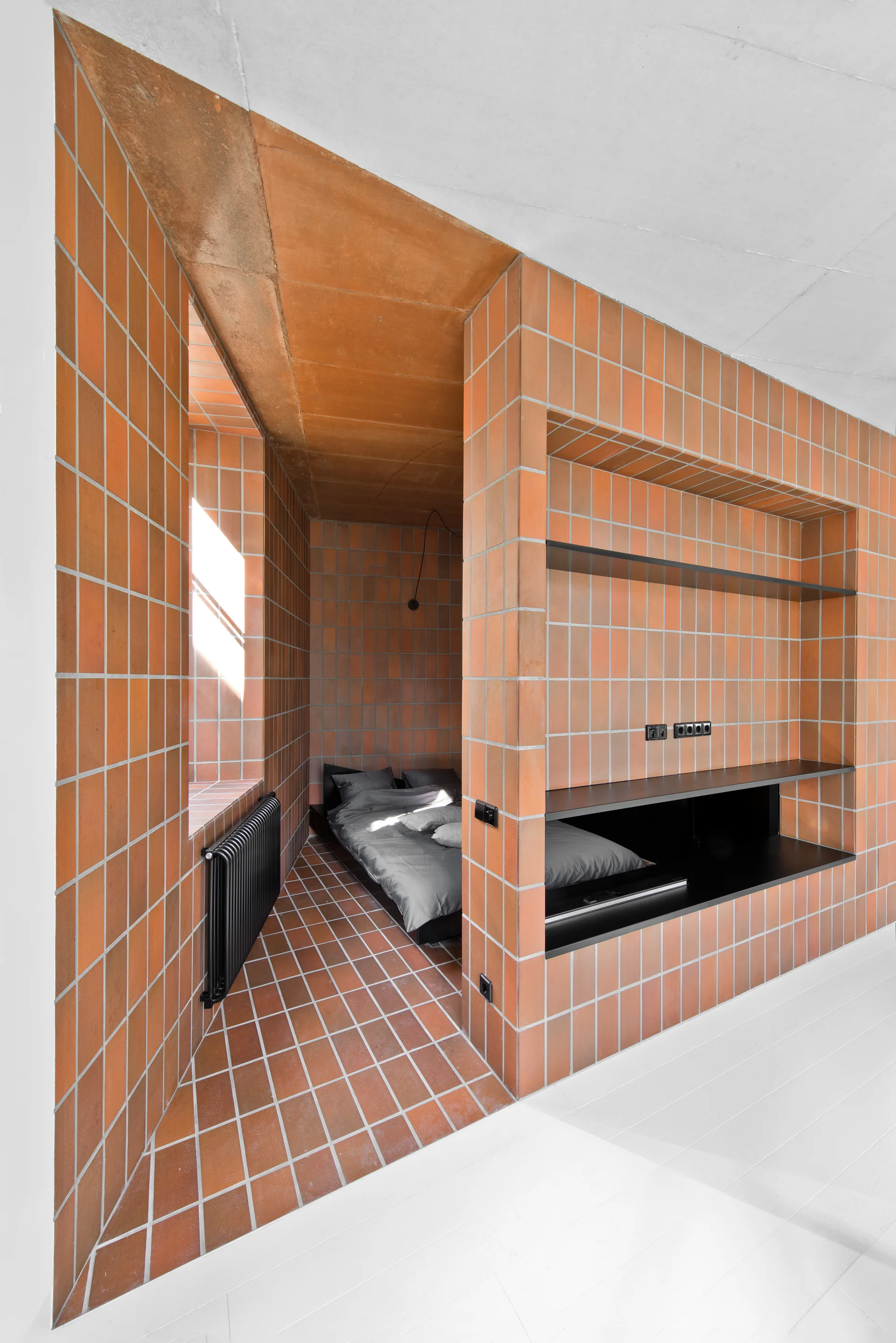 A minimalist bedroom featuring a sleeping alcove lined with orange terracotta tiles, a platform bed with grey linens, and black shelving.