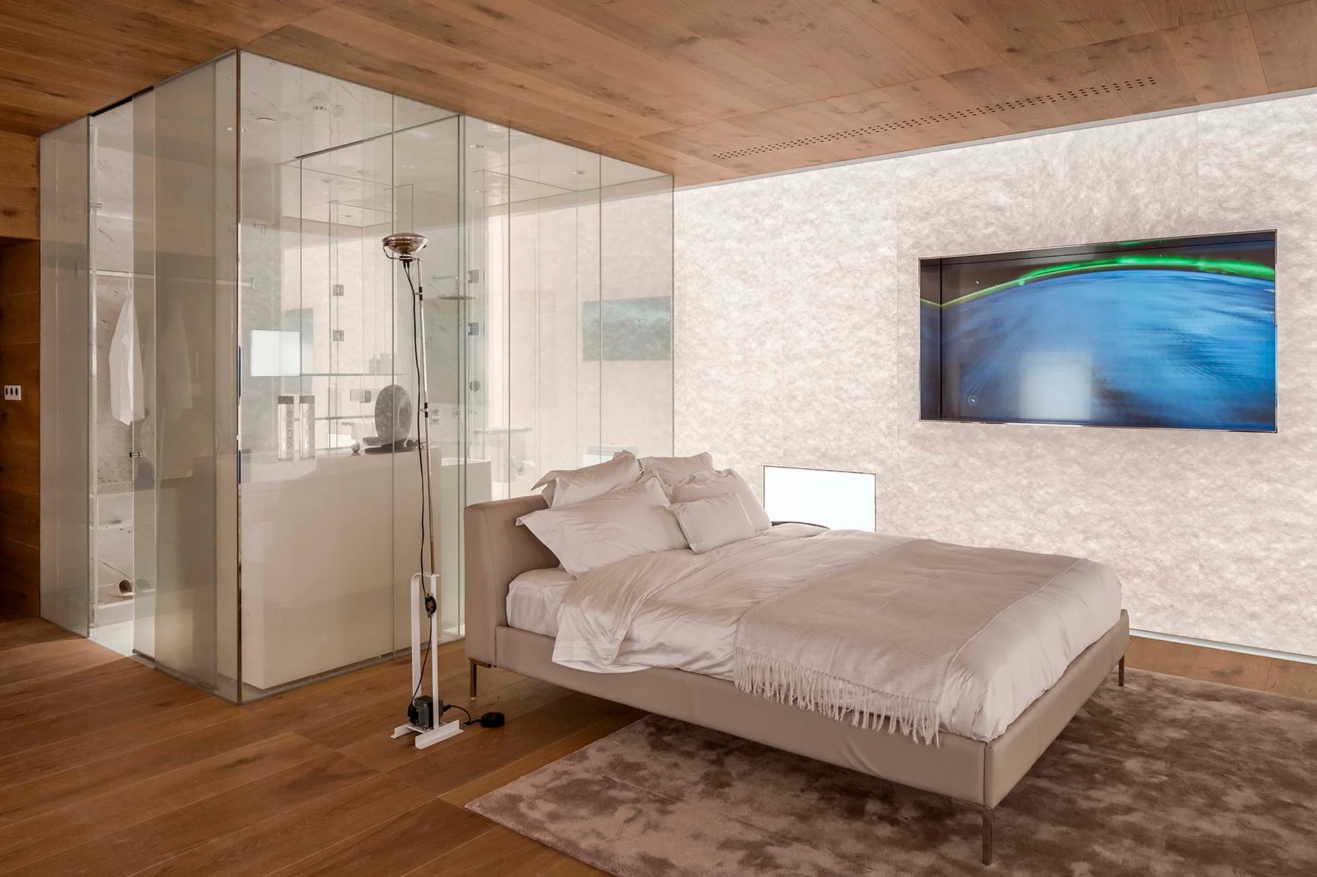 A contemporary bedroom features an upholstered bed, a glowing textured feature wall, and a transparent glass-enclosed bathroom.