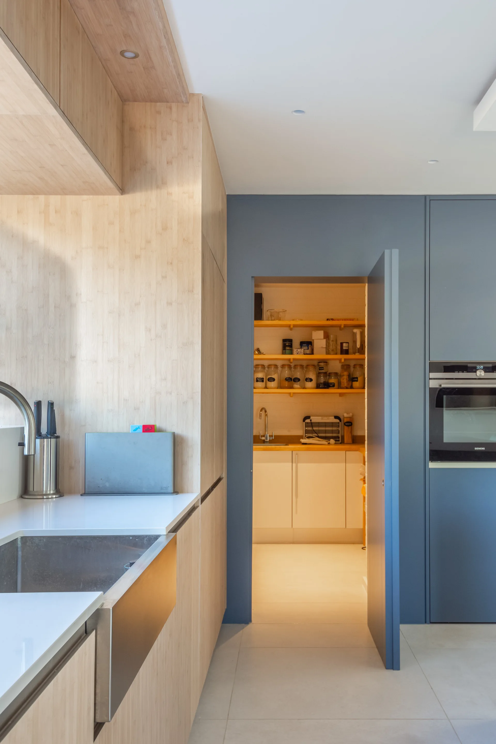 A modern kitchen featuring light wood cabinets, a stainless steel sink, and a matte blue wall with an open doorway leading to a walk-in pantry.