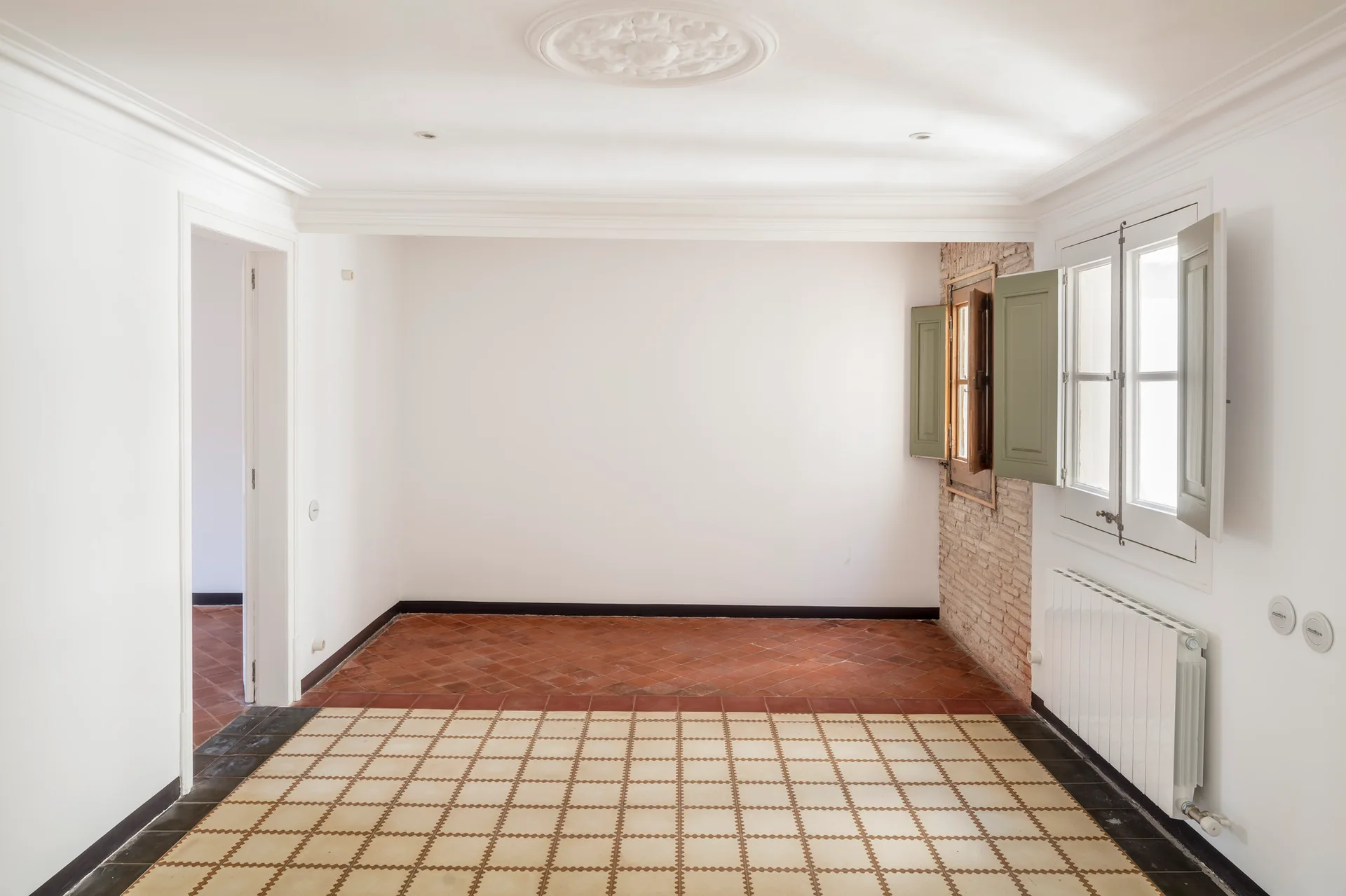 An empty room features a split floor of patterned and terracotta tiles, white walls with crown molding, and a window with sage green shutters.