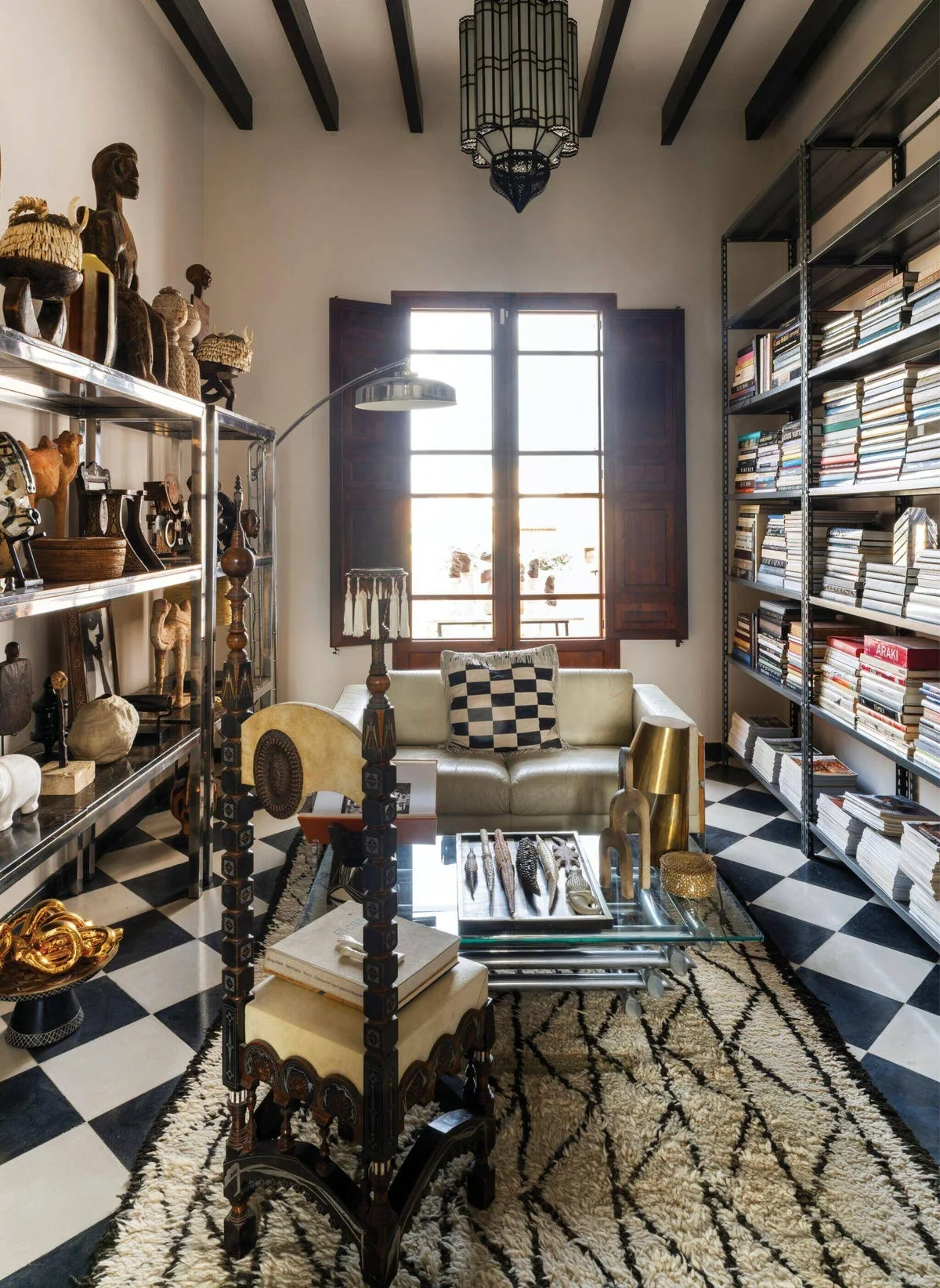 An eclectic library featuring tall metal shelves filled with books and collectibles, a cream leather sofa, and a black-and-white checkered floor.