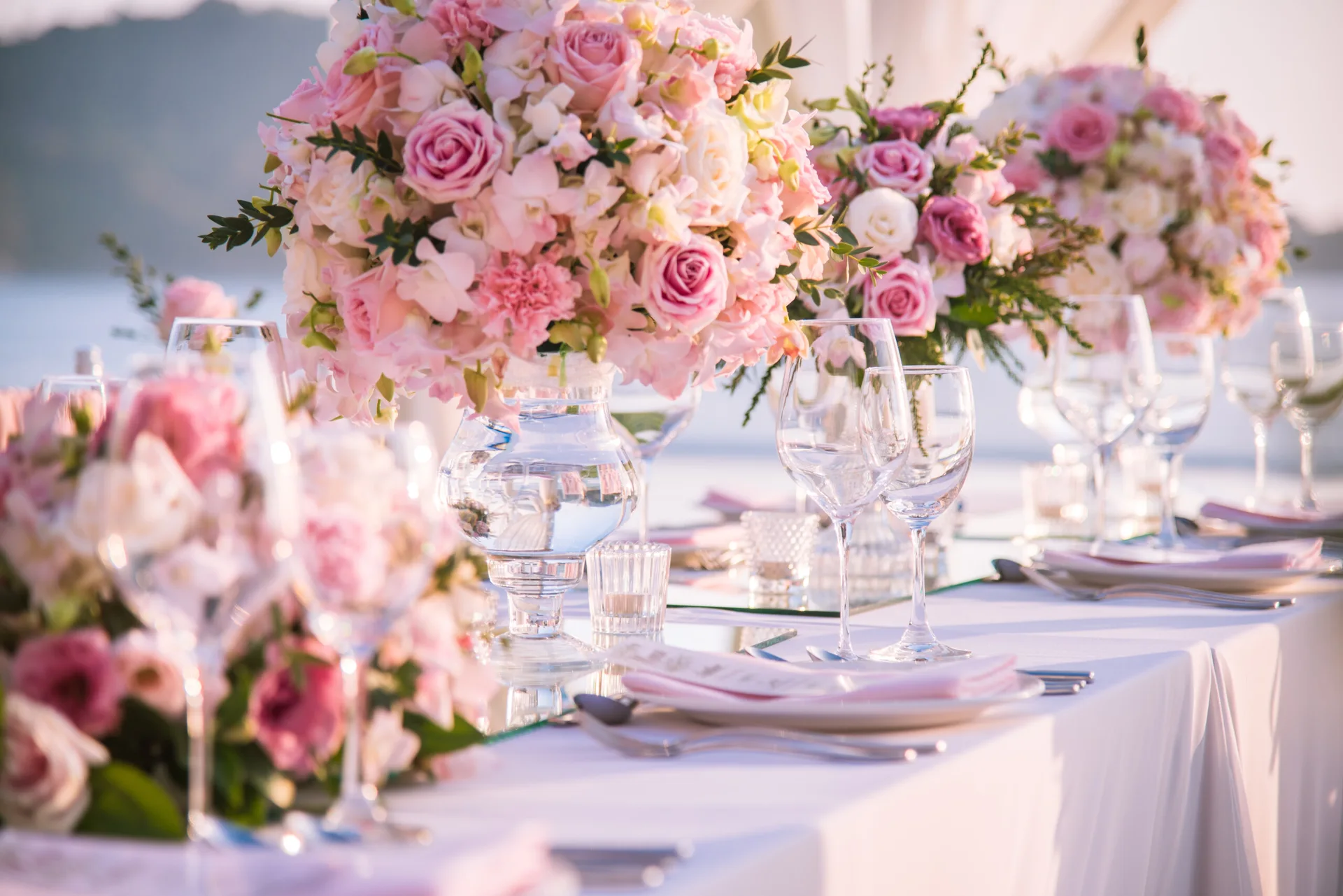 A long banquet table set with white linens, clear wine glasses, pink napkins, and large pink rose centerpieces on mirrored bases.