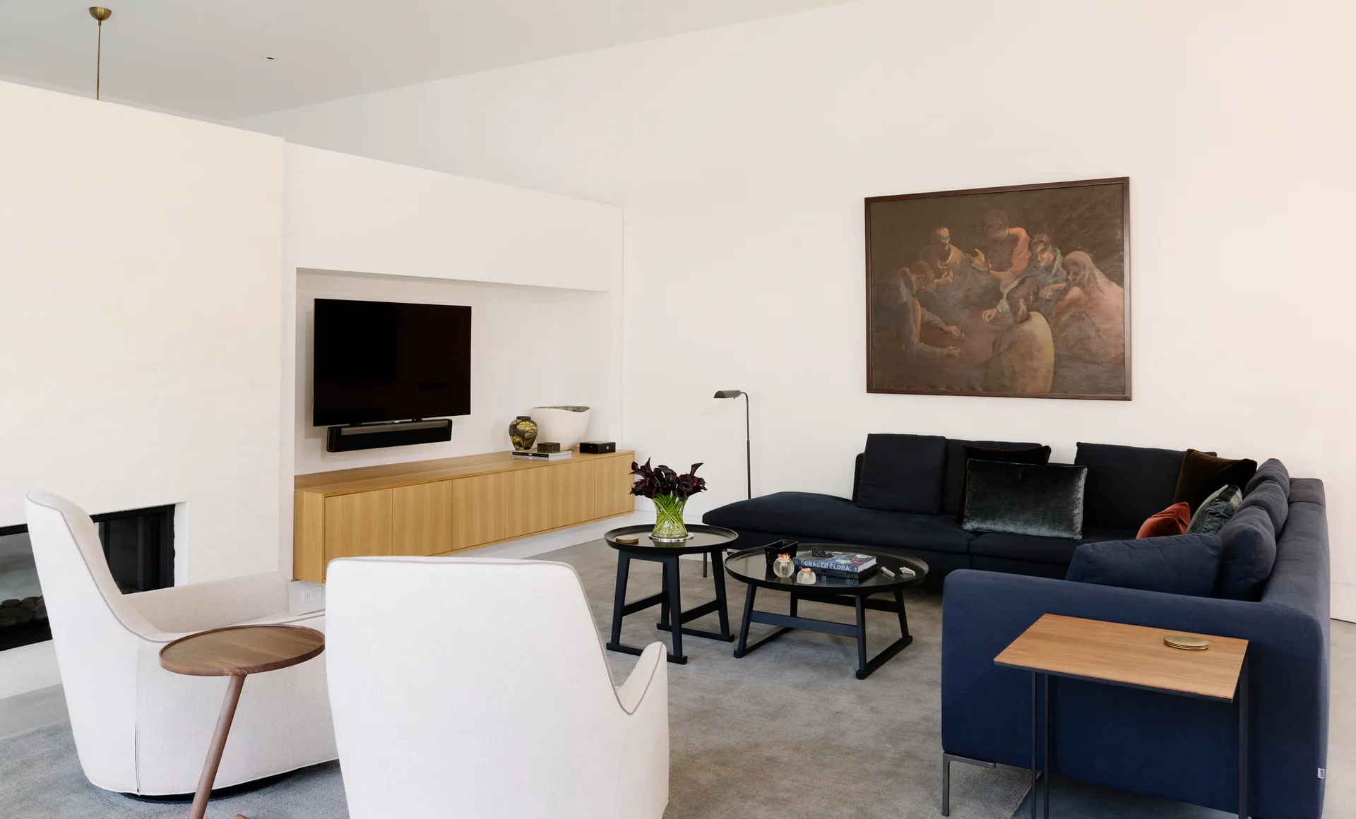 A minimalist living room features a navy blue sectional sofa, two off-white armchairs, and a floating wood console beneath a recessed television.