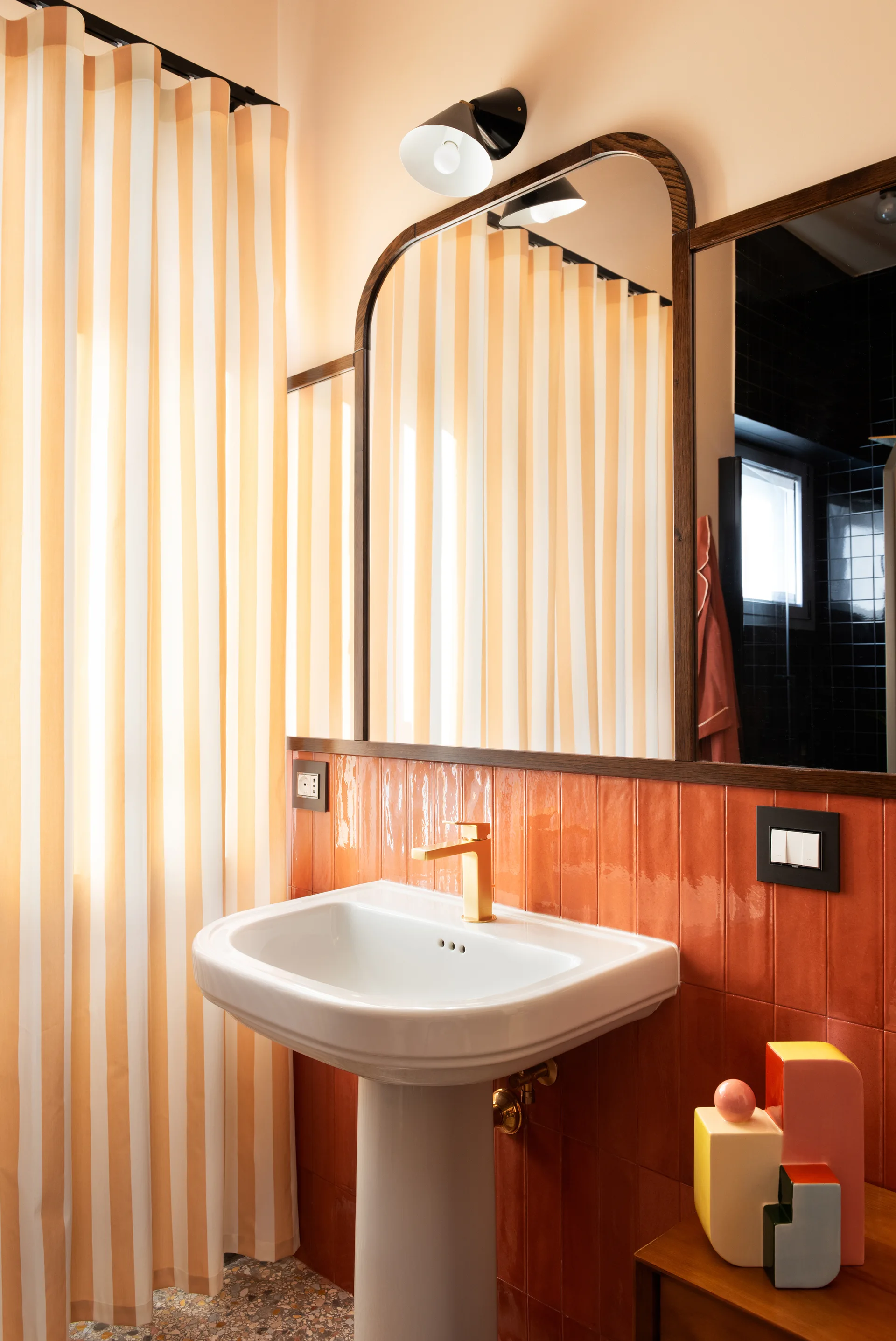 A bathroom vanity featuring a white pedestal sink against glossy terracotta wall tiles, alongside a striped curtain and an arched wood-framed mirror.